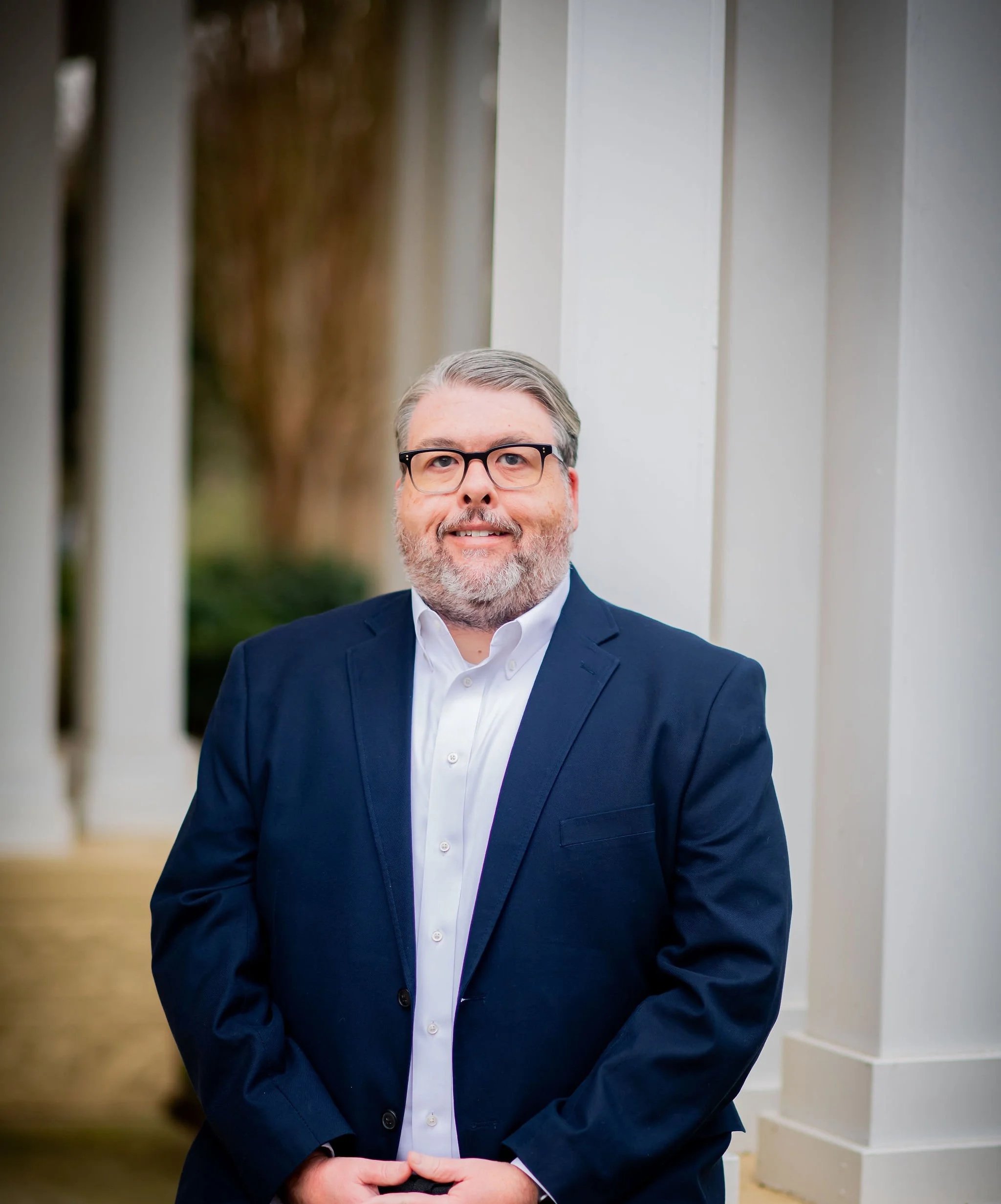 A man in a navy blue suit and white shirt standing outdoors, smiling in front of a white columned building with greenery and trees in the background.