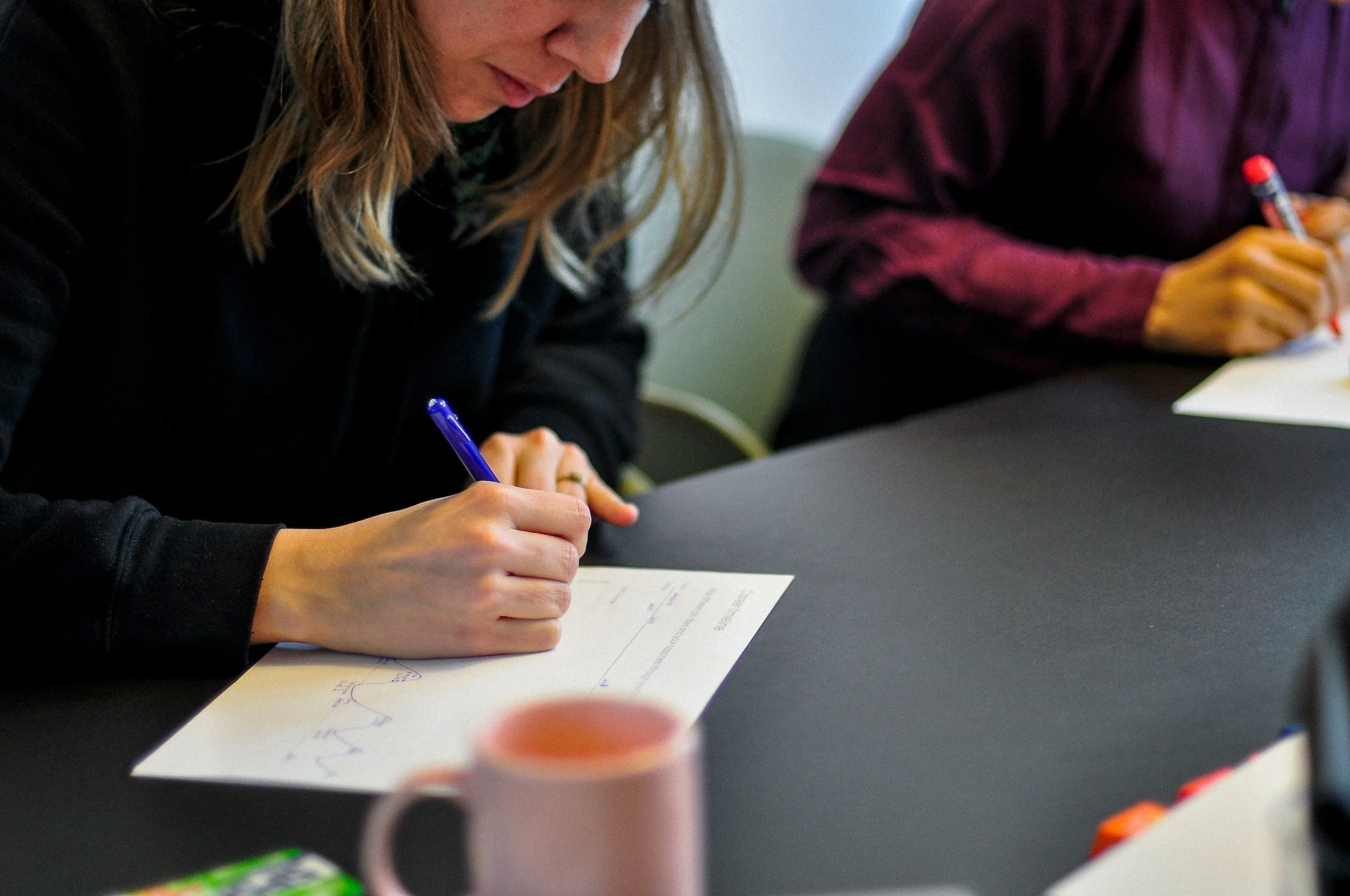 A woman sitting at a table, writing on a sheet of paper with a blue pen, with a pink mug in front of her. Another person in a maroon shirt also writing at the same table.