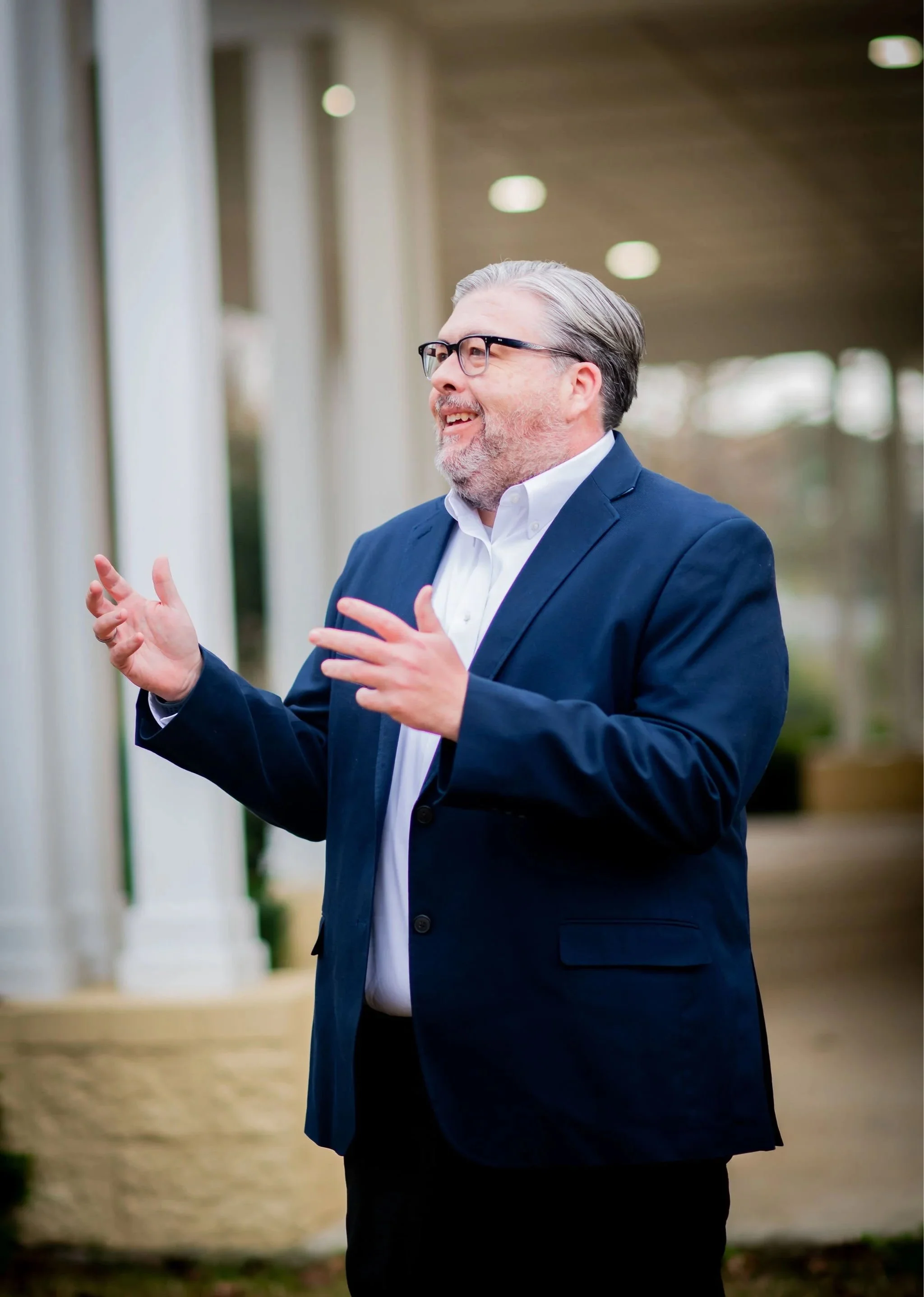 A man with glasses and gray hair, dressed in a navy suit and white shirt, is gesturing with his hands and smiling outdoors near a building with white columns.