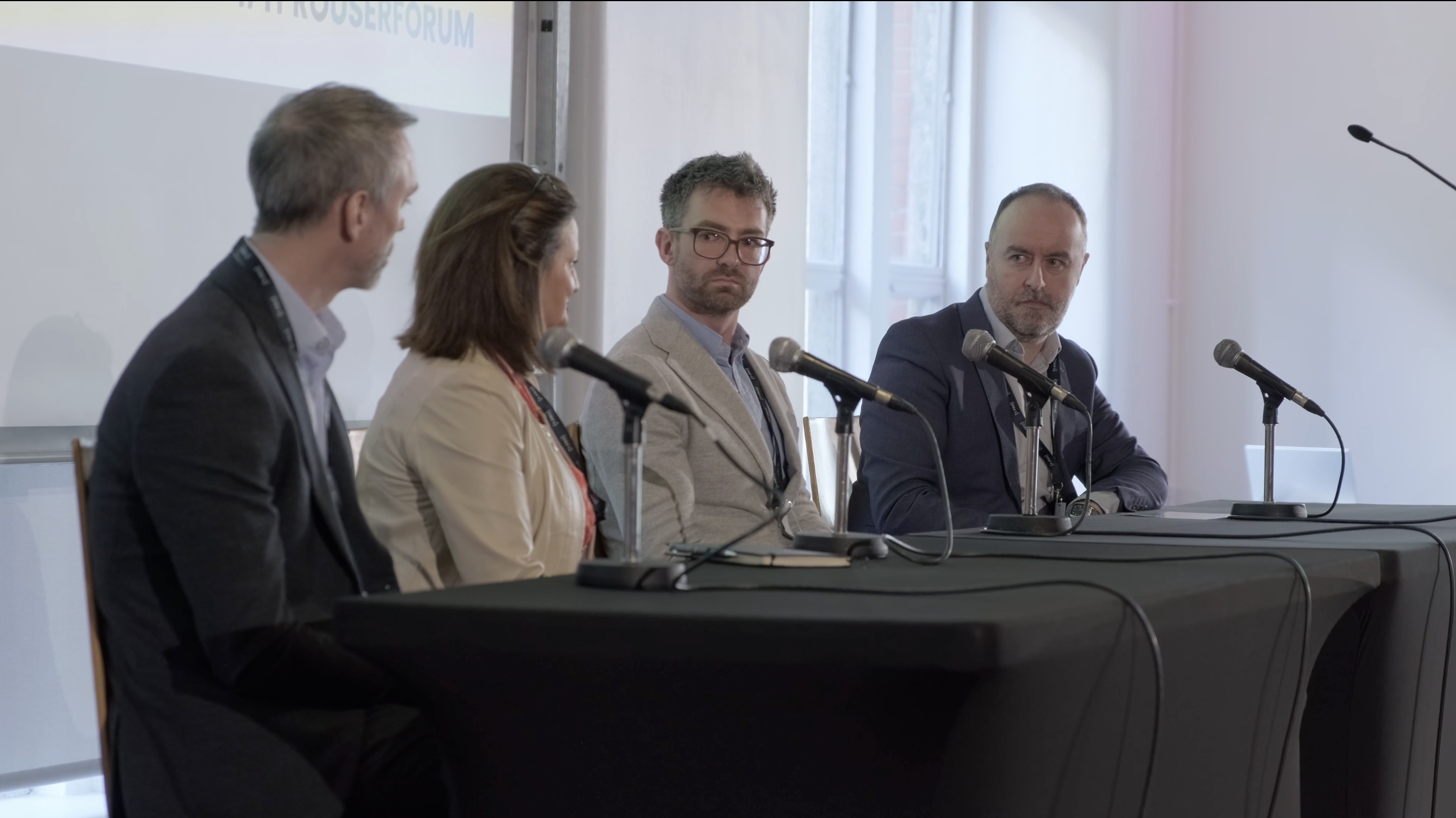 Four people seated at a panel table during a conference or panel discussion, with microphones in front of them, in a well-lit room with large windows.