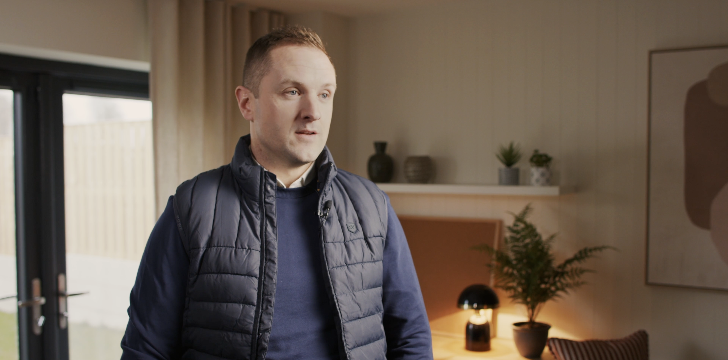 Man with short hair wearing a navy blue jacket and sweater standing inside a modern living room.