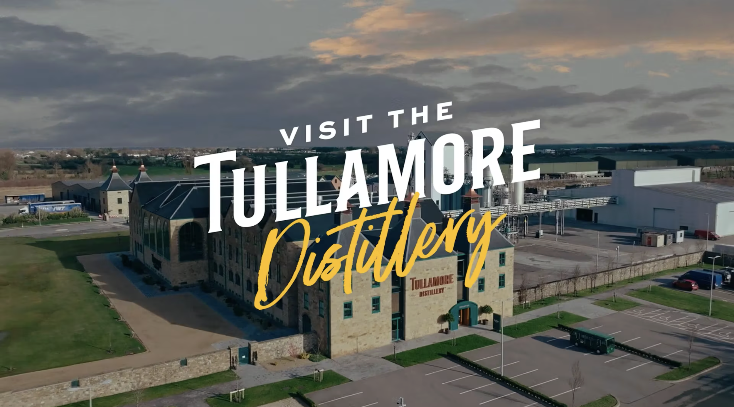 Aerial view of Tullamore Distillery building with parking lot in foreground and factory structures in background, under cloudy sky, with text overlay promoting a visit to Tullamore Distillery.