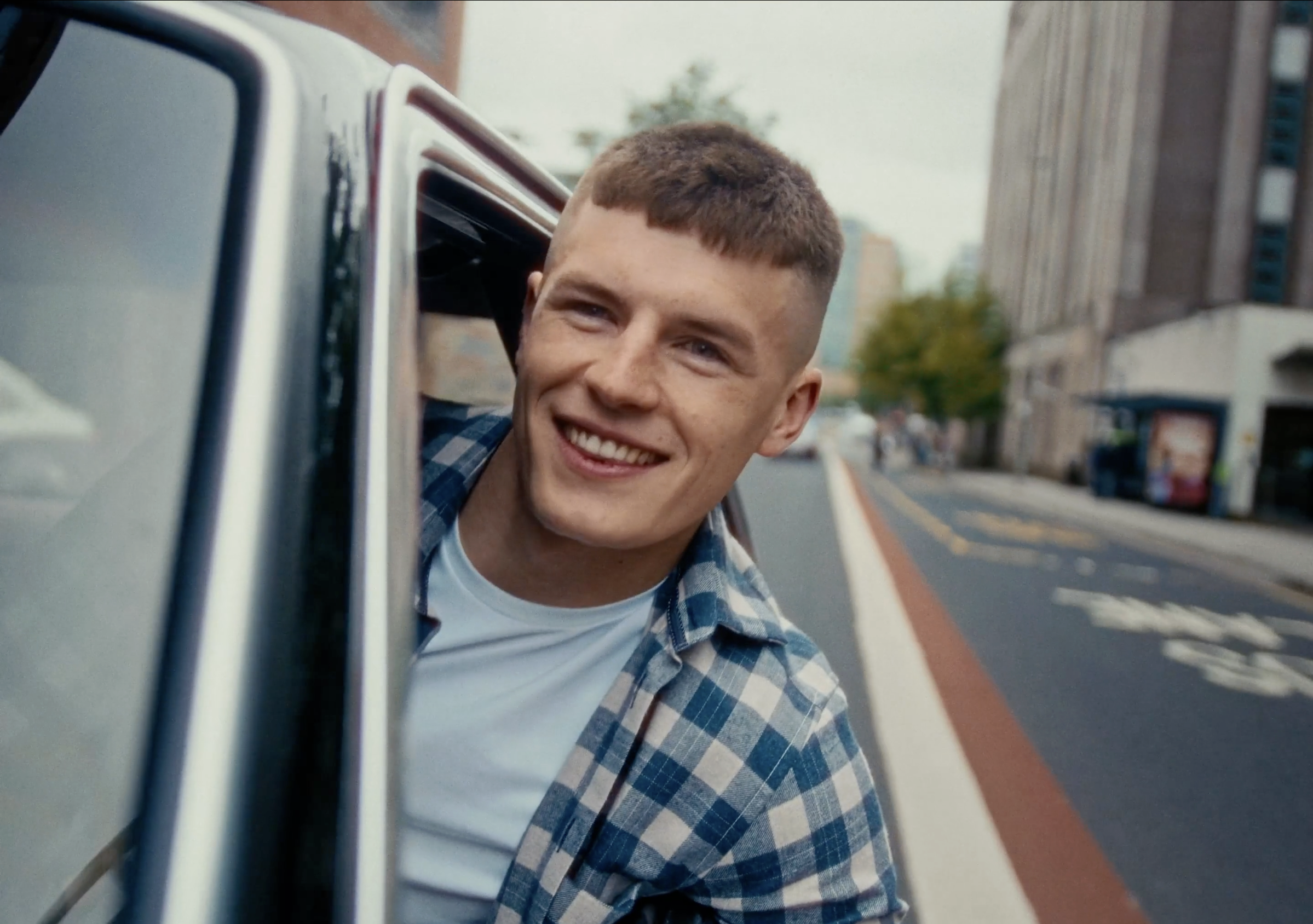 A young man with short brown hair and a plaid shirt leaning out of a car window, smiling on a city street.