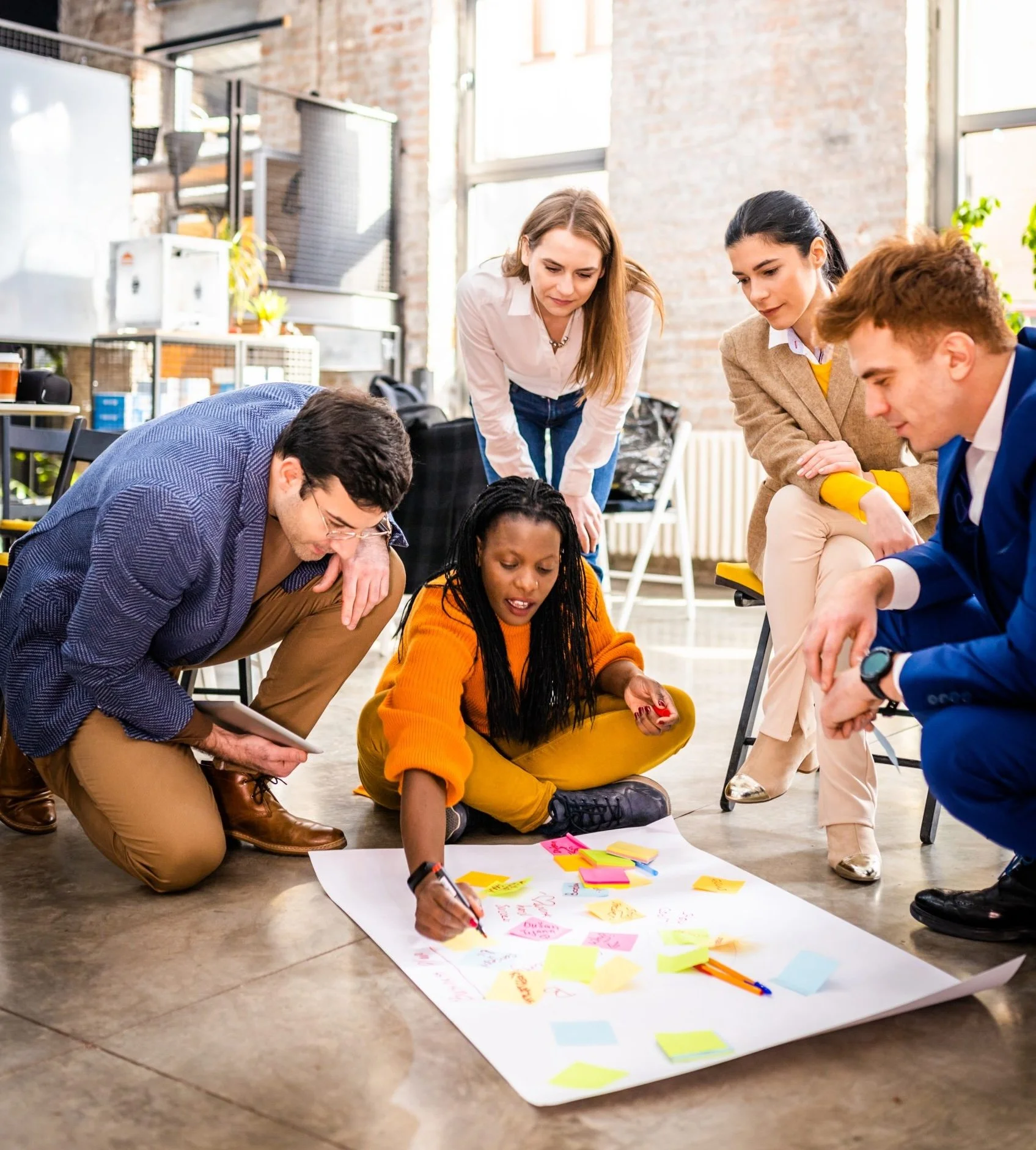Group of six diverse young adults working together on a project, sitting and kneeling around a large sheet of paper with colorful sticky notes and drawings, in a bright, industrial-style office space.