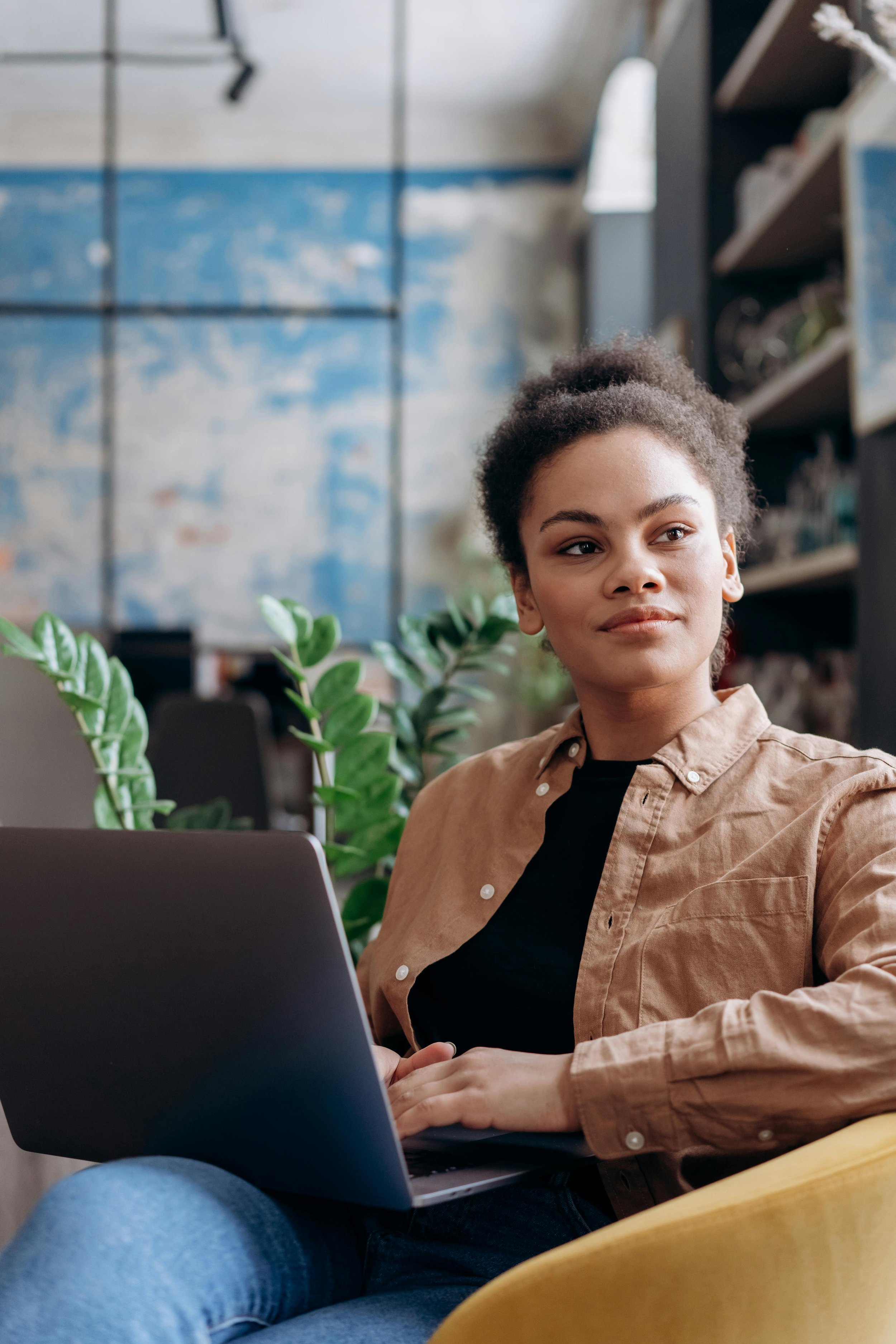 A woman with curly hair sitting on a yellow chair with a laptop on her lap, surrounded by green plants in an indoor setting with a blue and white wall.