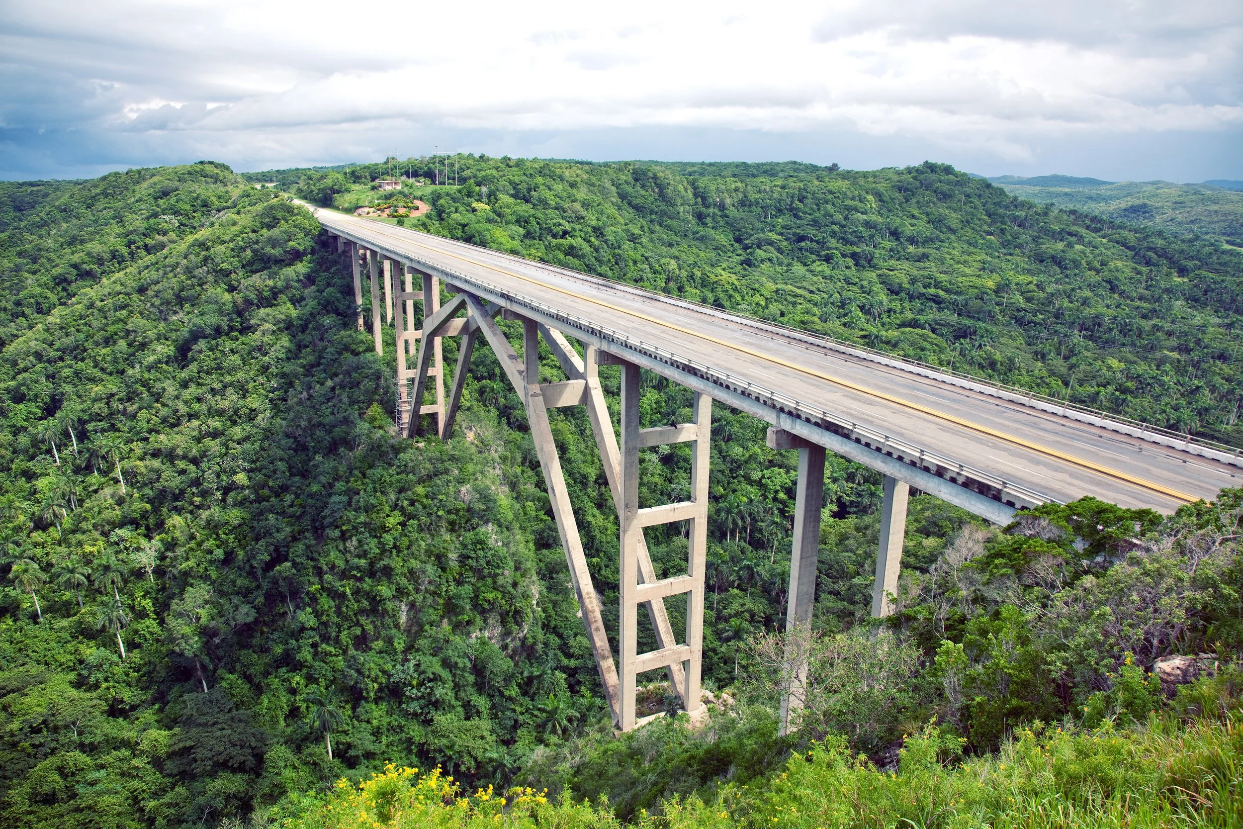 A large bridge stretching over a lush green valley with forested hills on either side, under a cloudy sky.