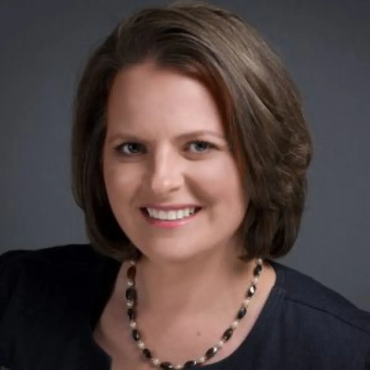 Close-up portrait of a woman with short, brown hair, smiling, wearing a black top and a black and pearl necklace, against a dark background.