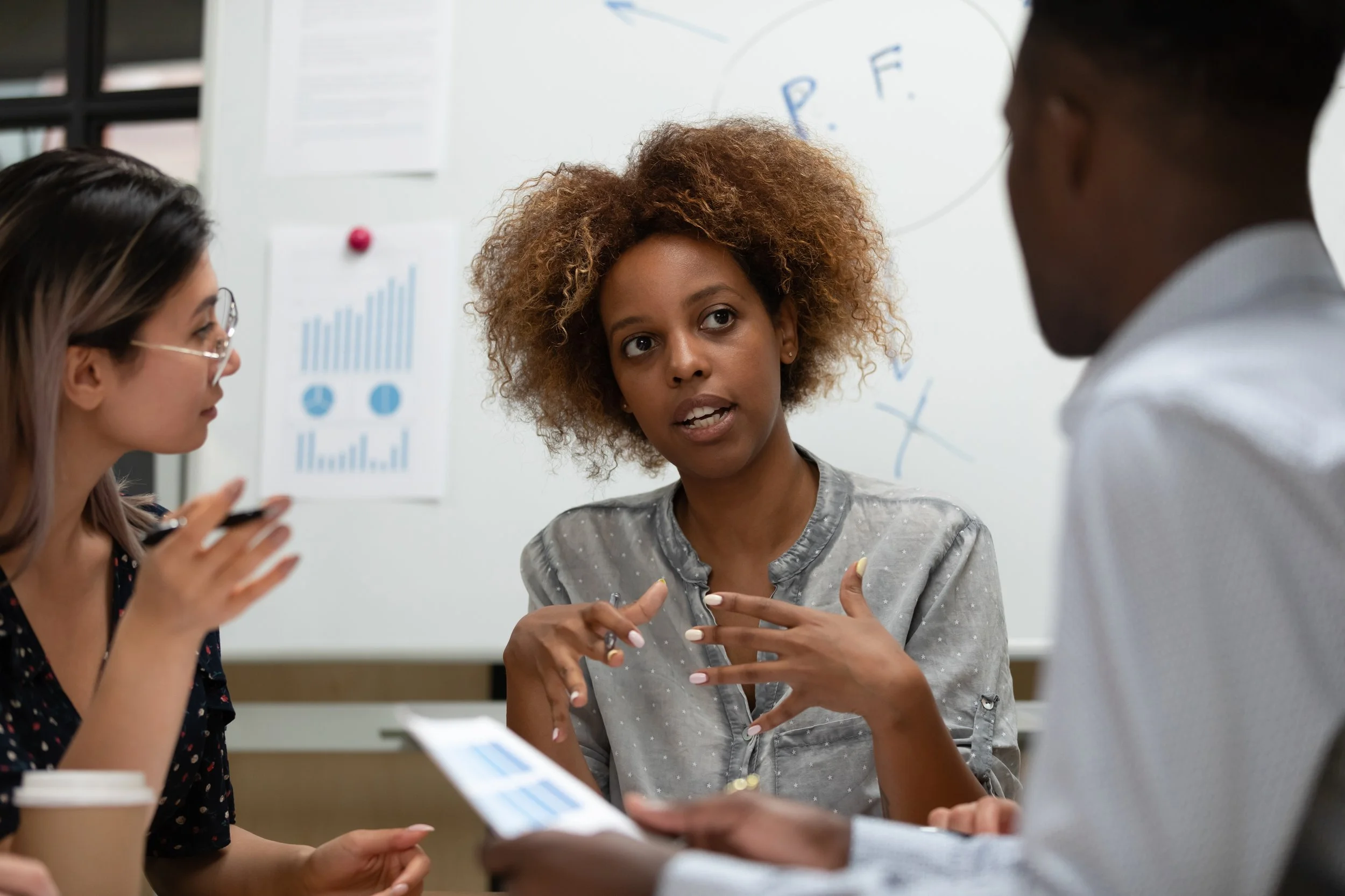 Three people engaged in a discussion in a conference room with charts and graphs on the wall and whiteboard behind them.