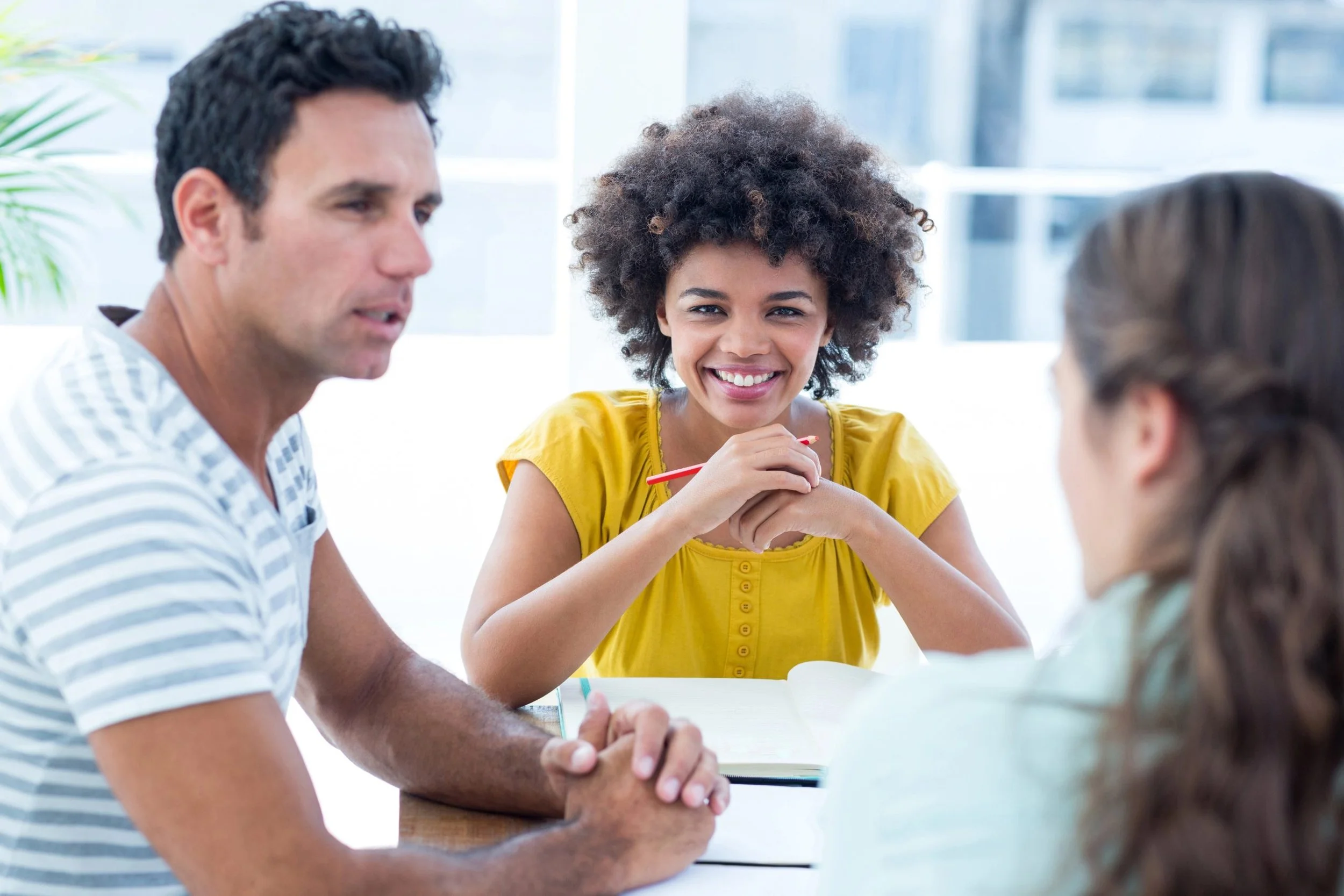 Four young adults sitting at a table, engaged in a discussion, with a woman smiling and holding a red pen, in a bright room with large windows.