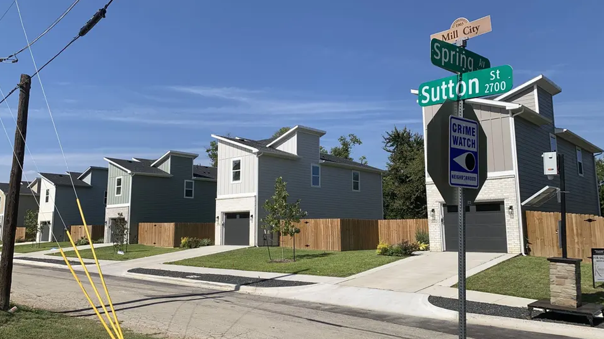 Newly built modern townhouses with gray exterior, small front lawns, and a wooden fence in a residential neighborhood under a clear blue sky. Street signs indicate the intersection of Spring Avenue, Sutton Street, and Mill City.