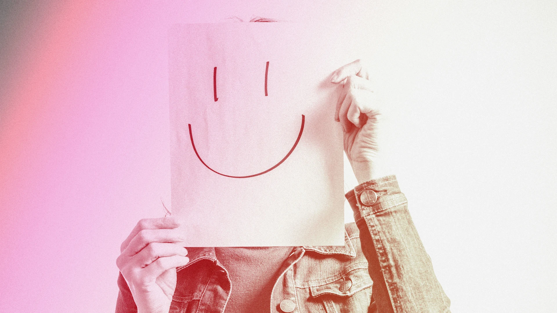 A person holding a piece of paper with a smiling face drawn on it, obscuring their face. The person is wearing a denim jacket and the background is light-colored.