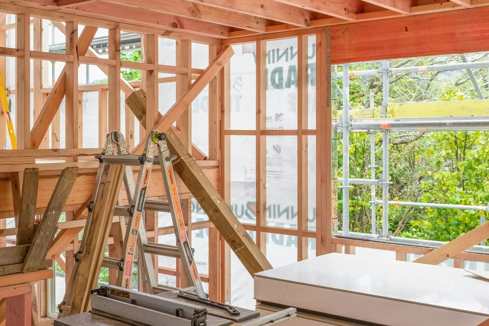 Interior view of a building under construction, showing wooden framing and scaffolding outside through the windows.