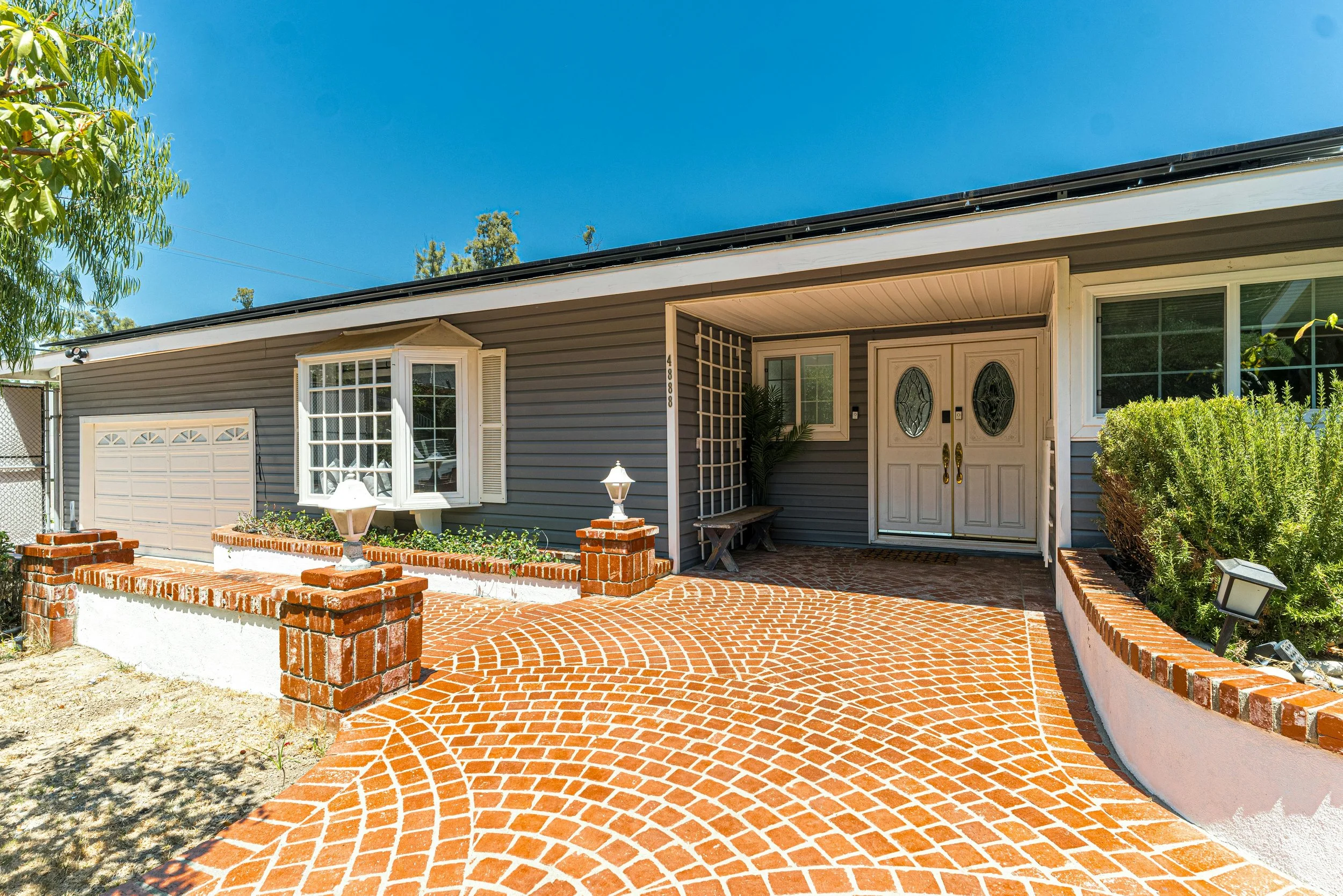 Front view of a house with a brick paver walkway, double front doors, a large window, shrubs, and a small garden area under a clear blue sky.