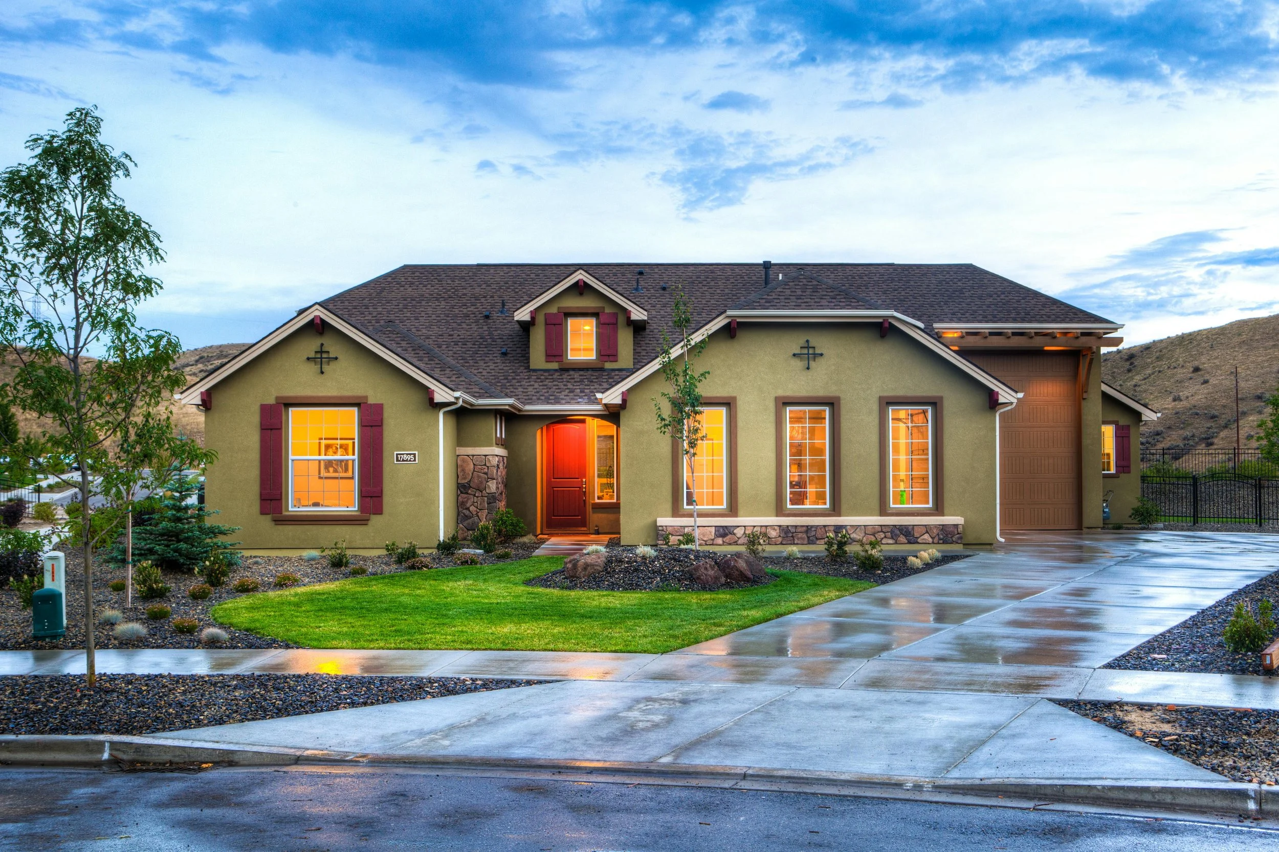 Front view of a modern house with green exterior, brown roof, and illuminated windows during dusk, with a wet driveway and landscaped yard.