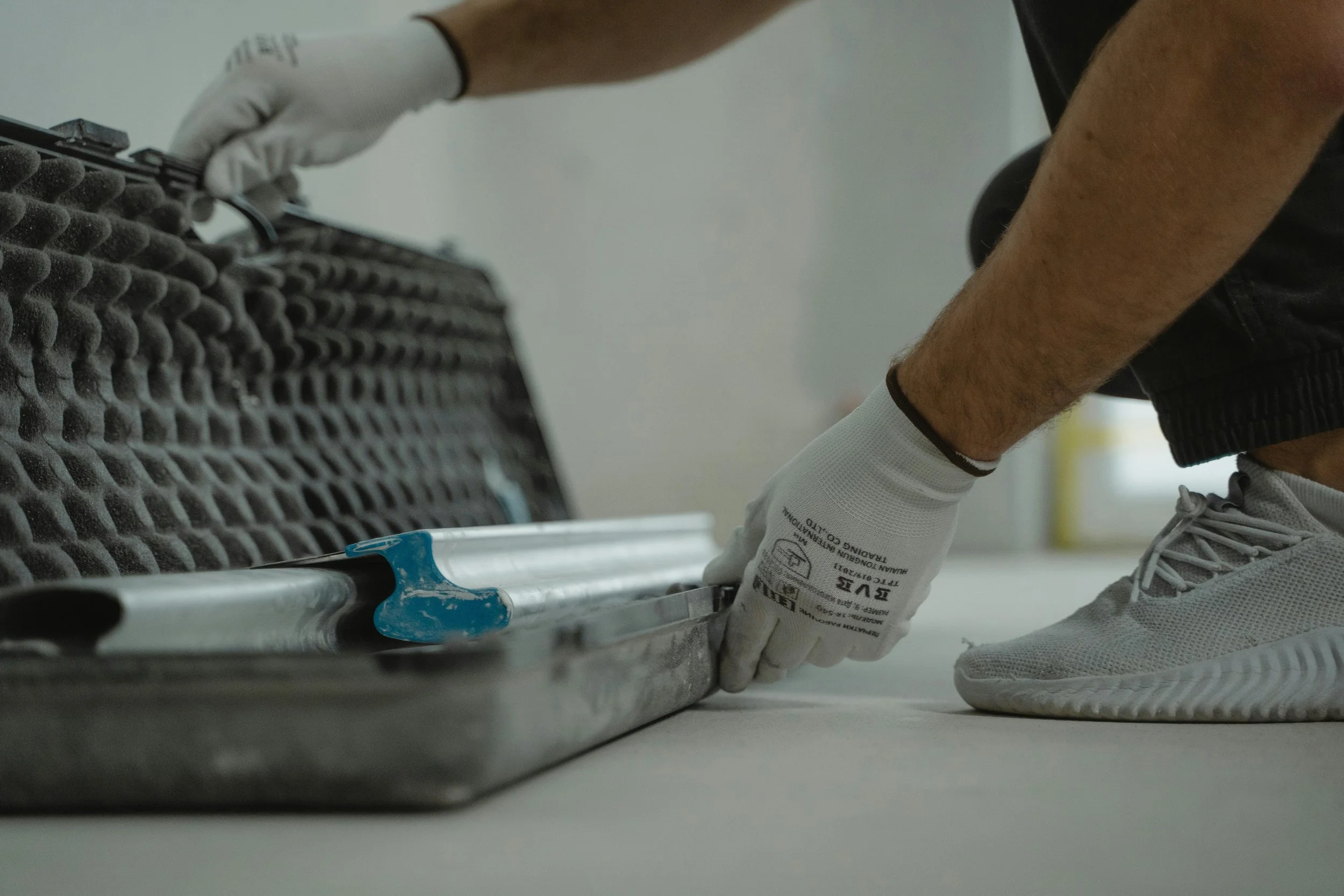 A person wearing gloves and athletic shoes is kneeling on the floor, handling a metal tray with foam padding on the side, possibly in a workshop or industrial setting.