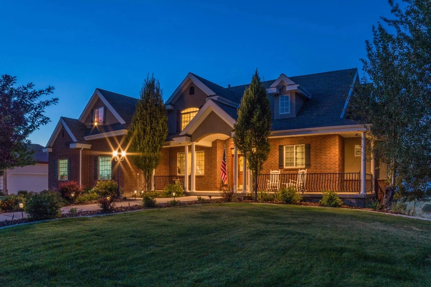A large brick house illuminated at dusk with a well-manicured lawn, tall trees, and outdoor lighting on the porch and yard.
