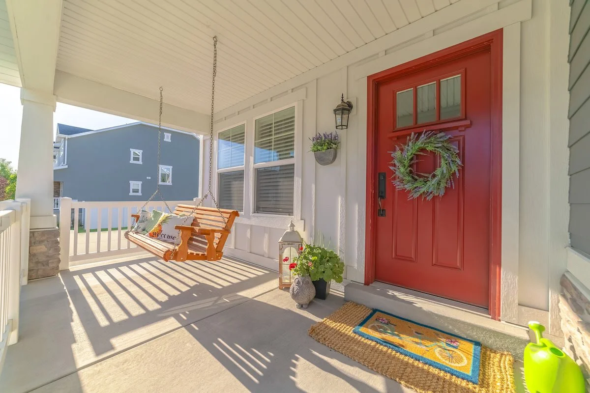 Front porch with a red front door adorned with a green wreath, a hanging wooden porch swing with cushions, potted plants, a lantern, a doormat, and a small green watering can.