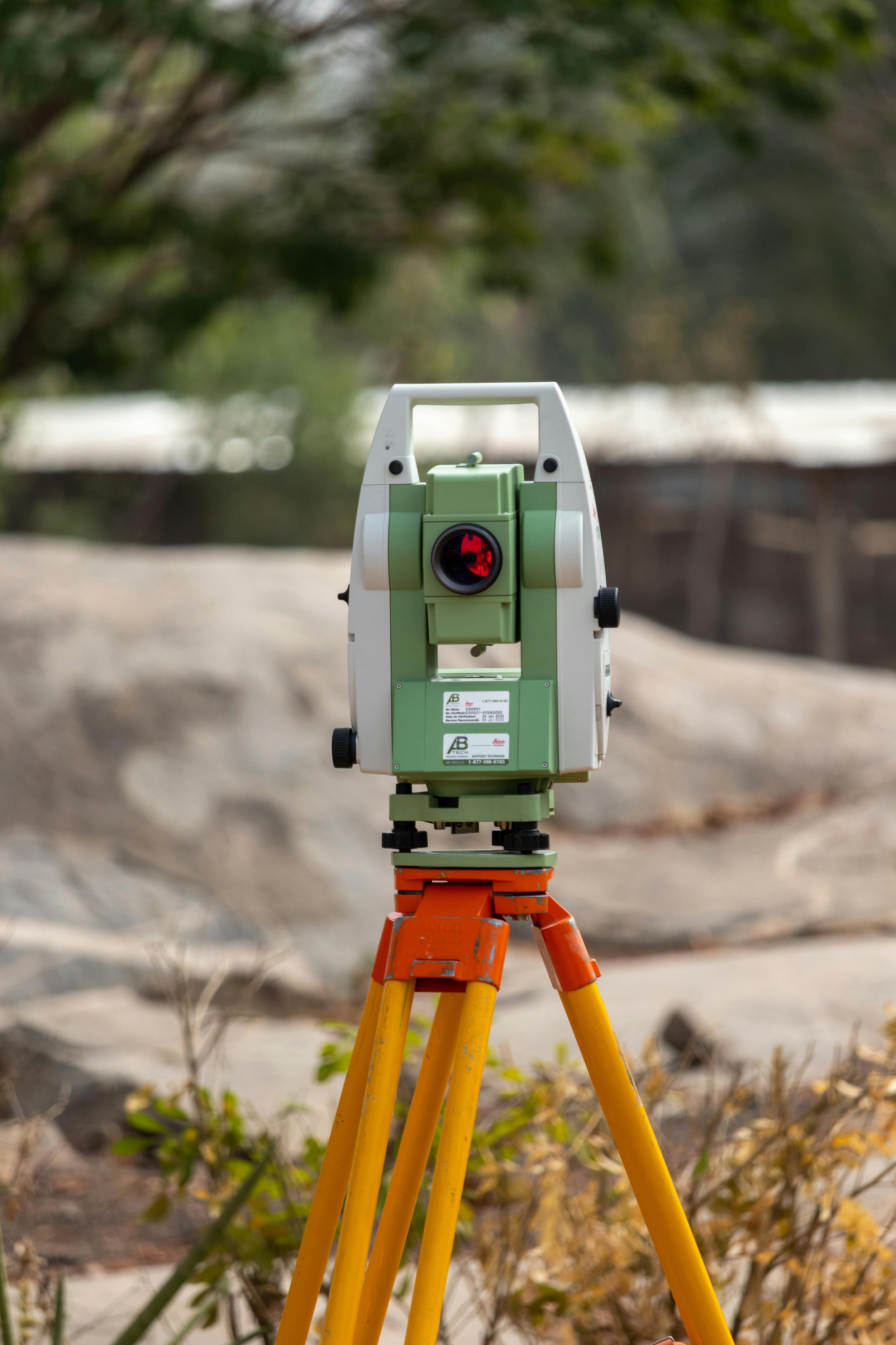 A total station surveying instrument set up outdoors on a tripod, with trees and a rocky terrain in the background.