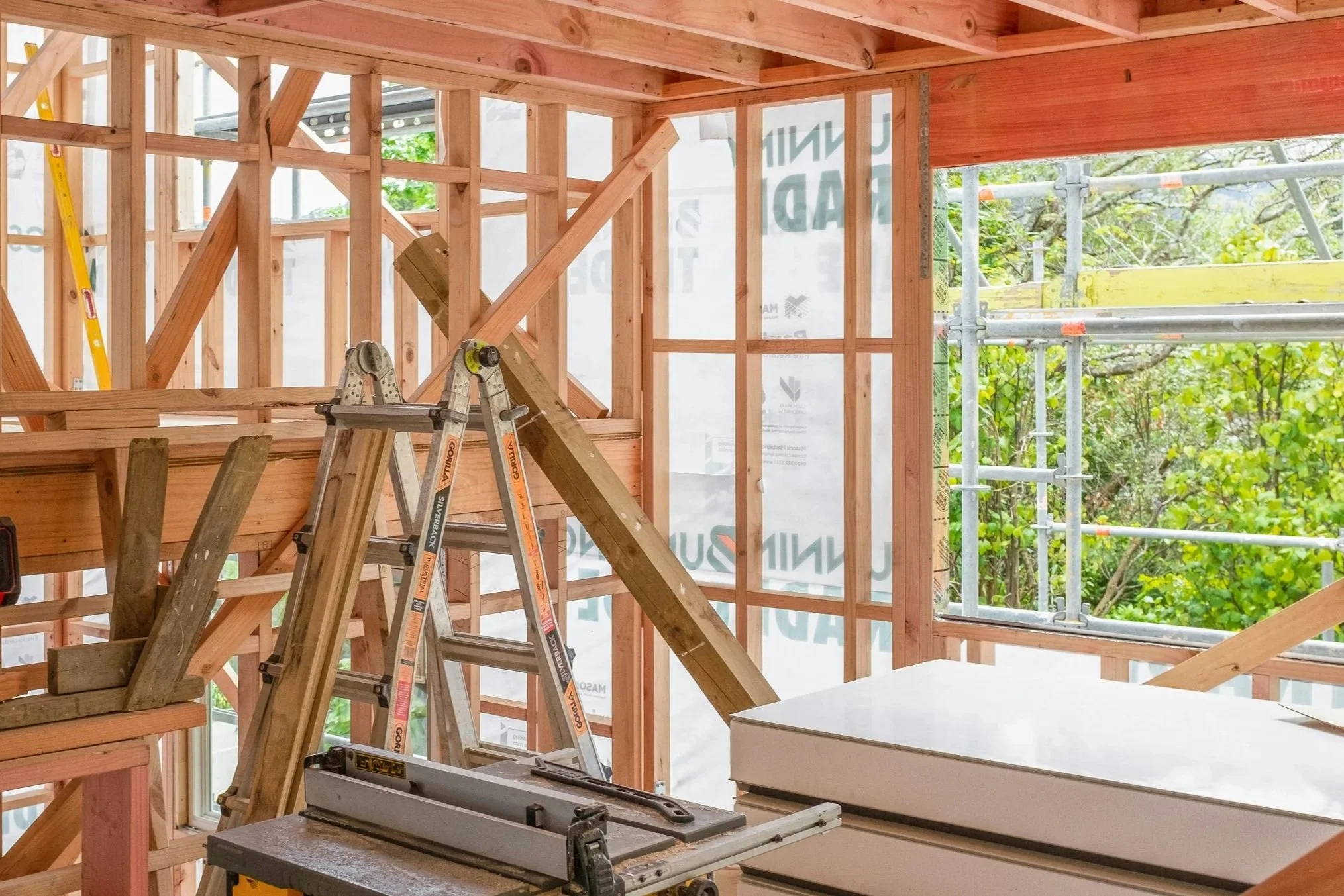 Inside a building under construction, with exposed wooden framing, a ladder, and scaffolding visible outside through the windows.
