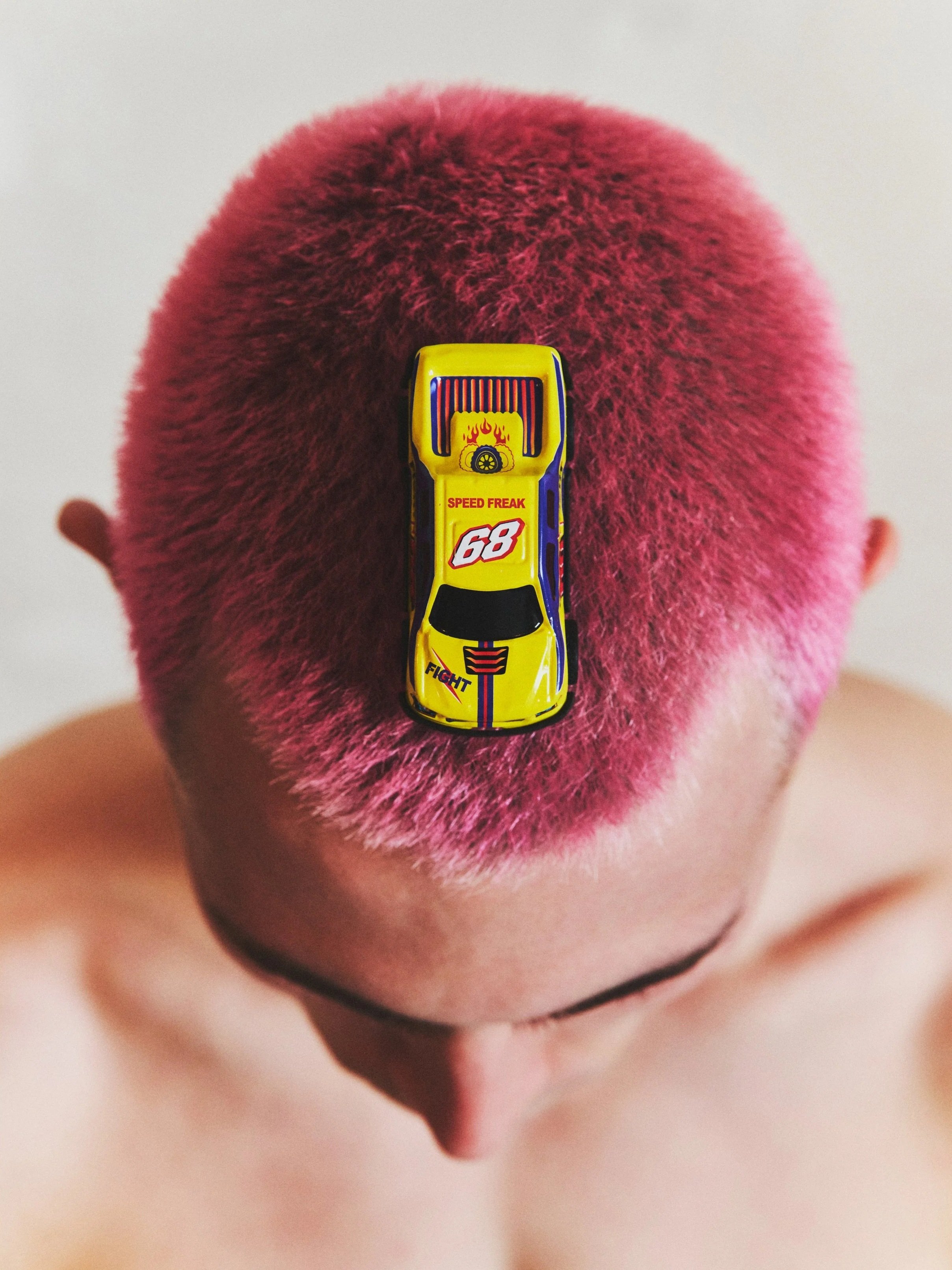 Persona con cabello rubio cortado muy corto, teñido de rosa brillante, con una chapa de coche de juguete en la parte superior de la cabeza.