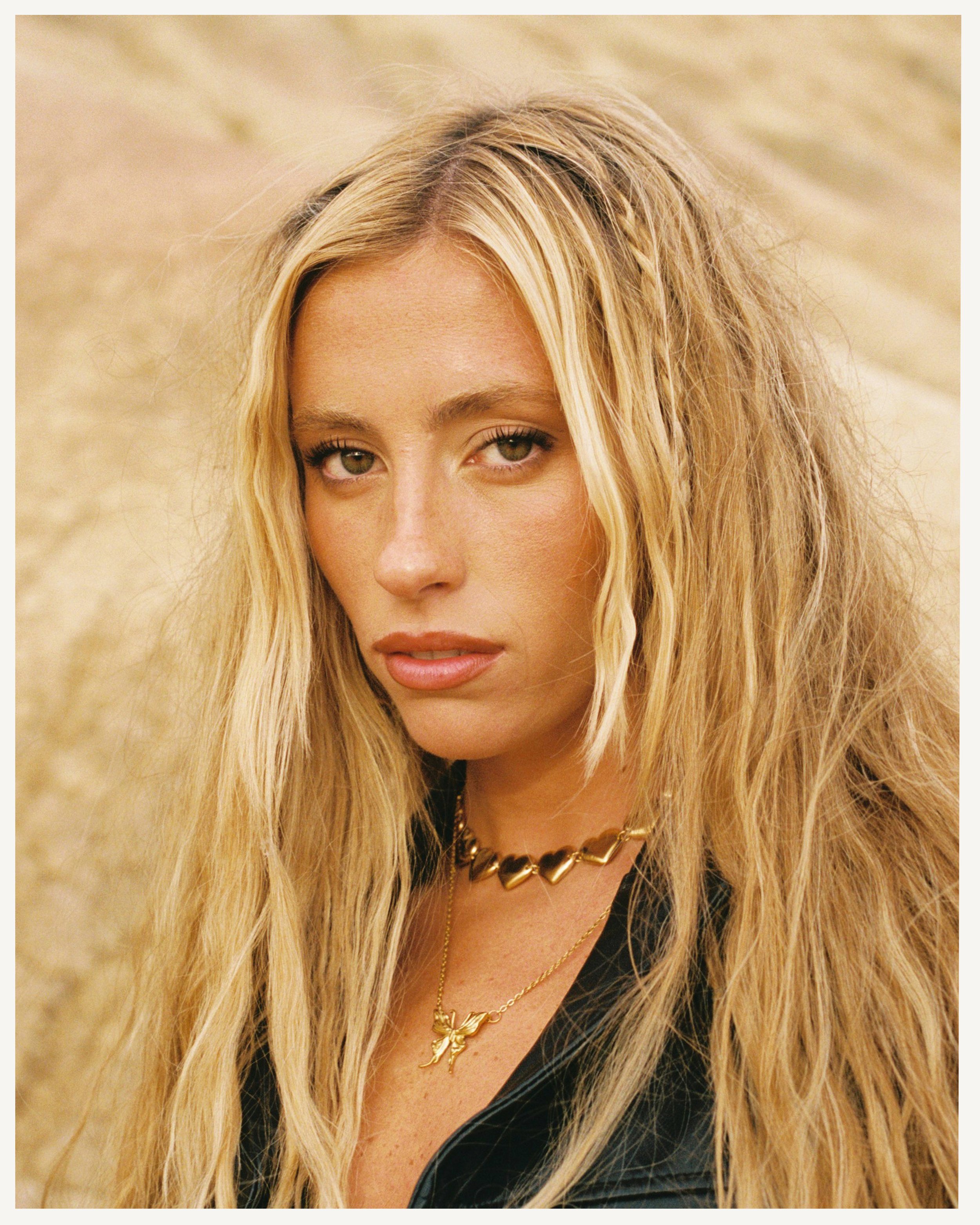 Retrato de una mujer con cabello rubio, ojos claros, usando collares dorados y una chaqueta negra, en un fondo arenoso.