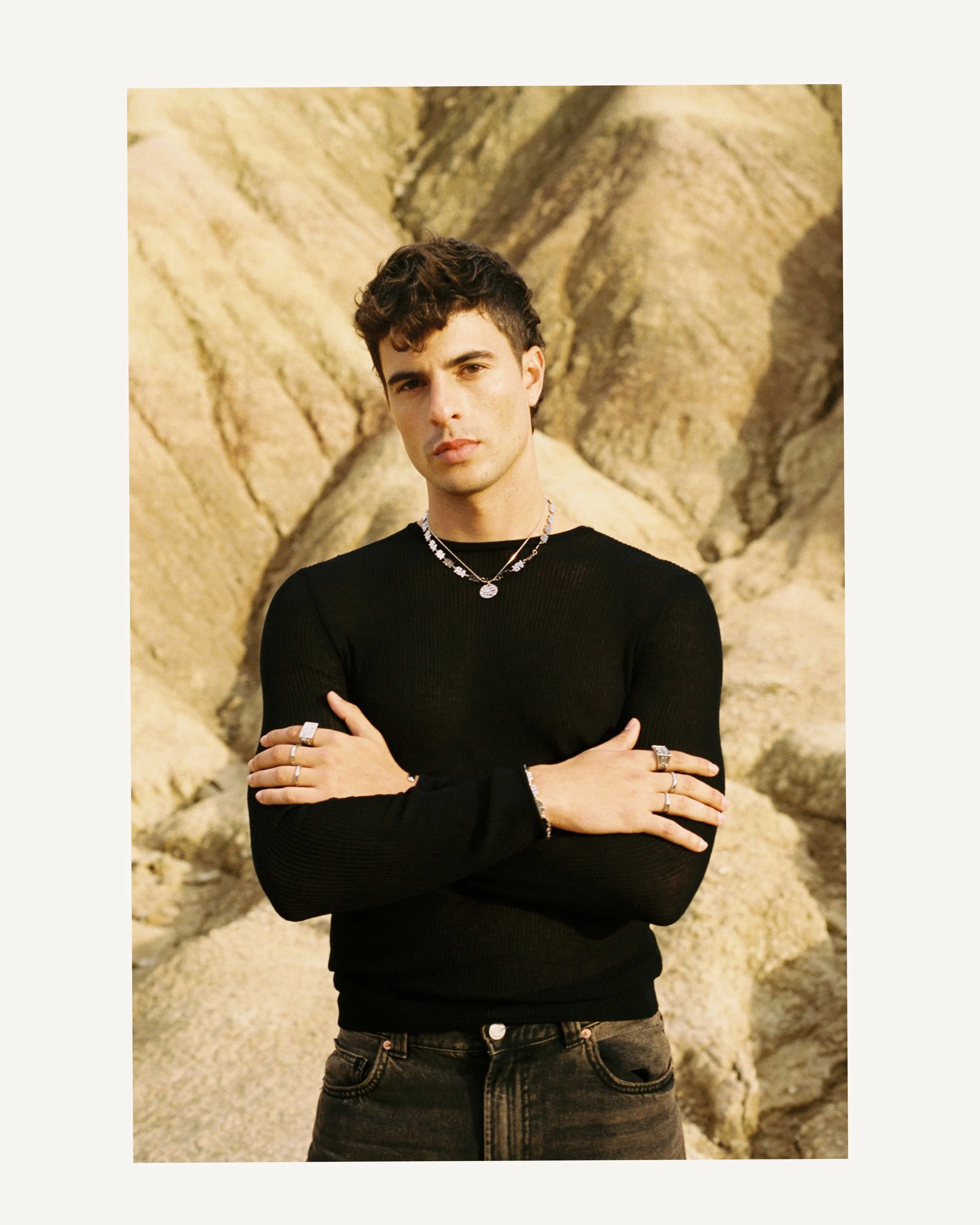 Joven con cabello rizado en un paisaje de rocas con expresión seria, usando ropa negra y accesorios de plata.