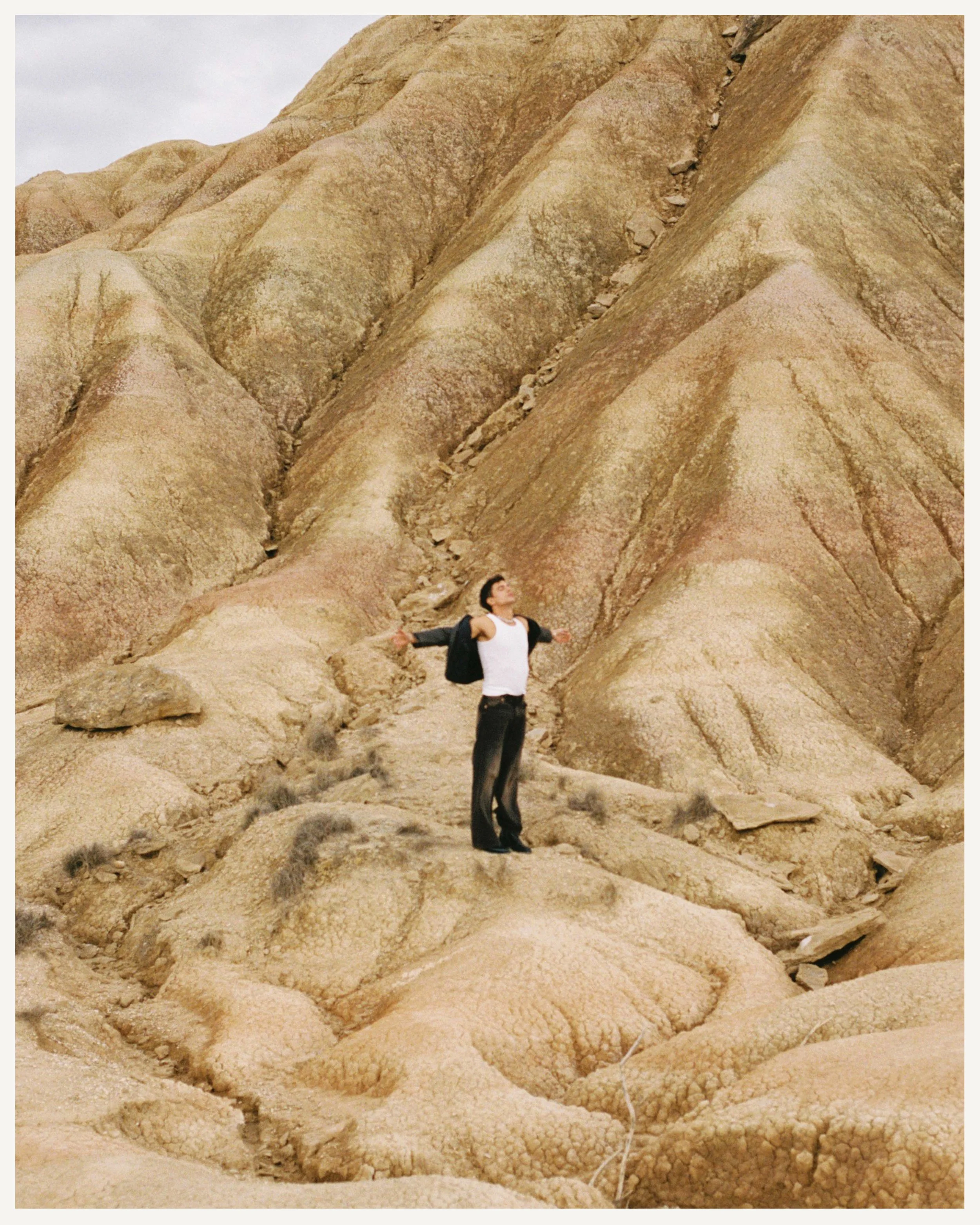 Persona con chaqueta y camiseta blanca en un paisaje árido con formaciones rocosas beige.