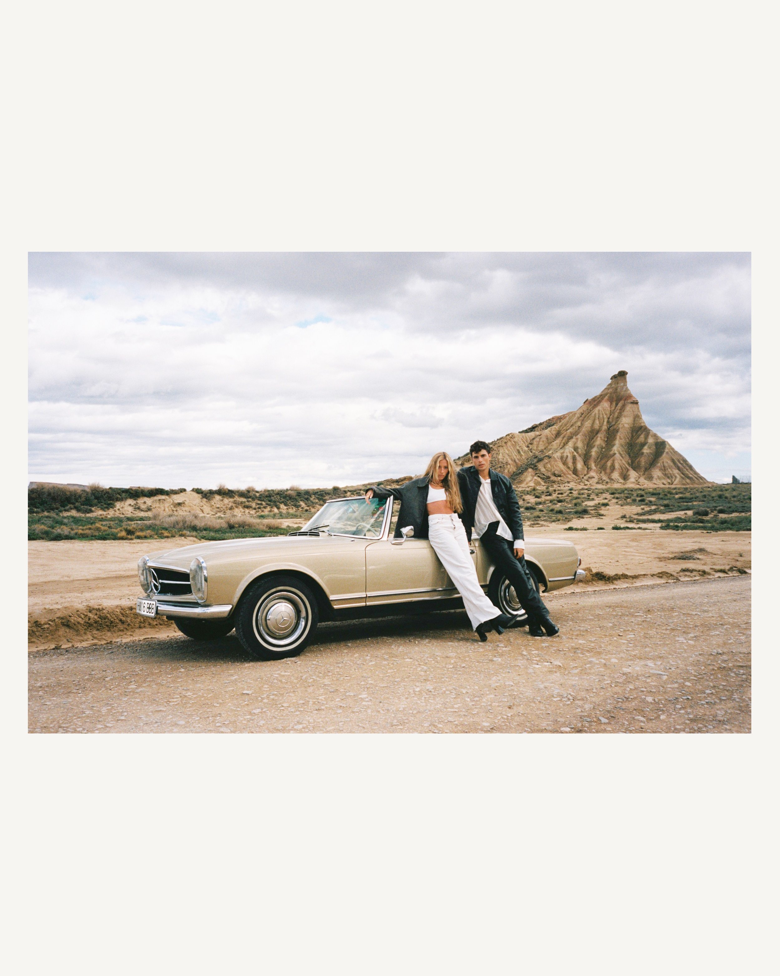 Dos personas adolescentes posando junto a un coche clásico en un paisaje desértico con una montaña en forma de pirámide en el fondo.
