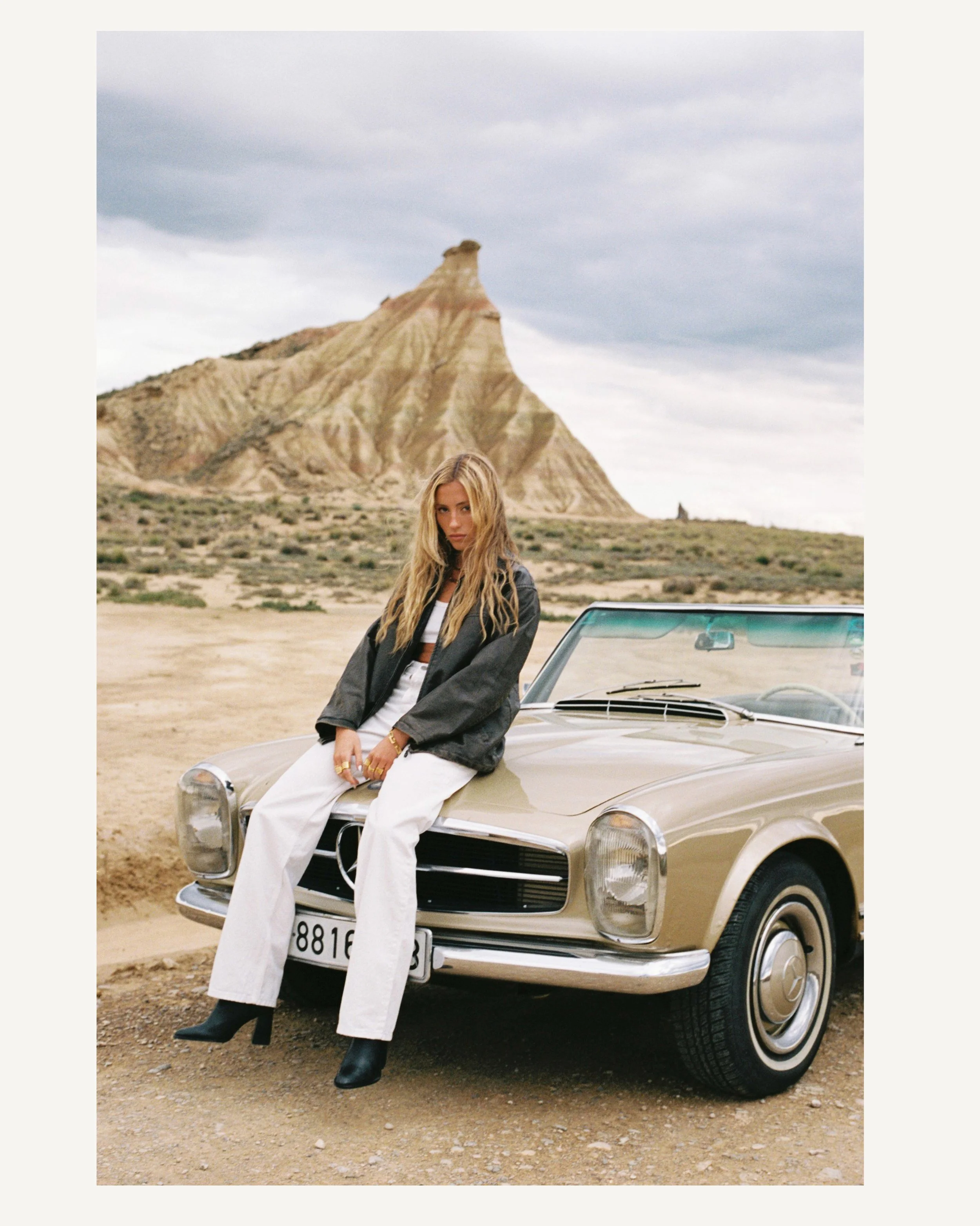 Una mujer con cabello rubio y largo, vestida con chaqueta negra y pantalones blancos, se apoya en un automóvil clásico de color beige en un paisaje desértico con montañas en el fondo y cielo nublado.