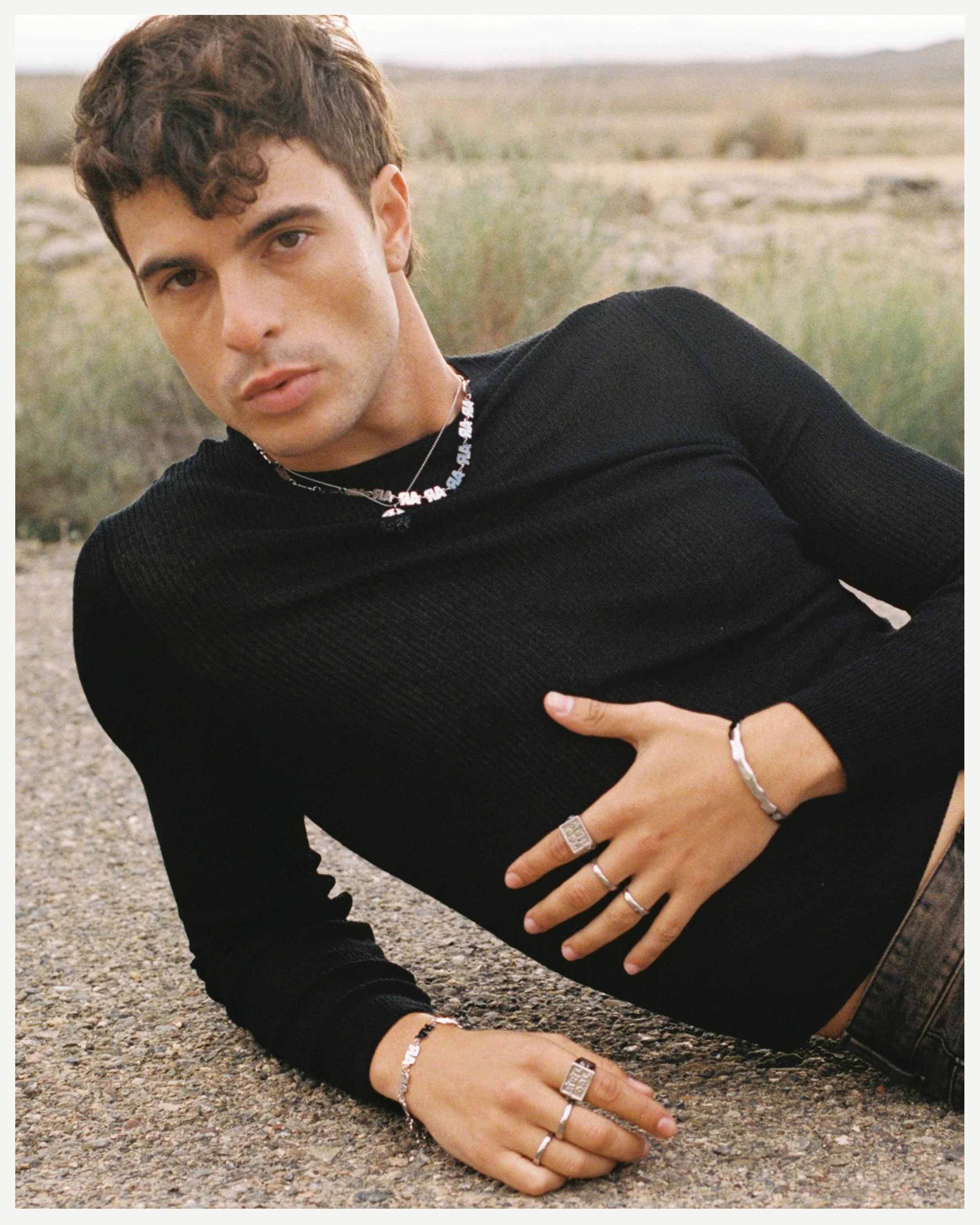 Un joven acostado en el suelo en un paisaje árido y natural, lleva puesta ropa de color negro y varias joyas plateadas.