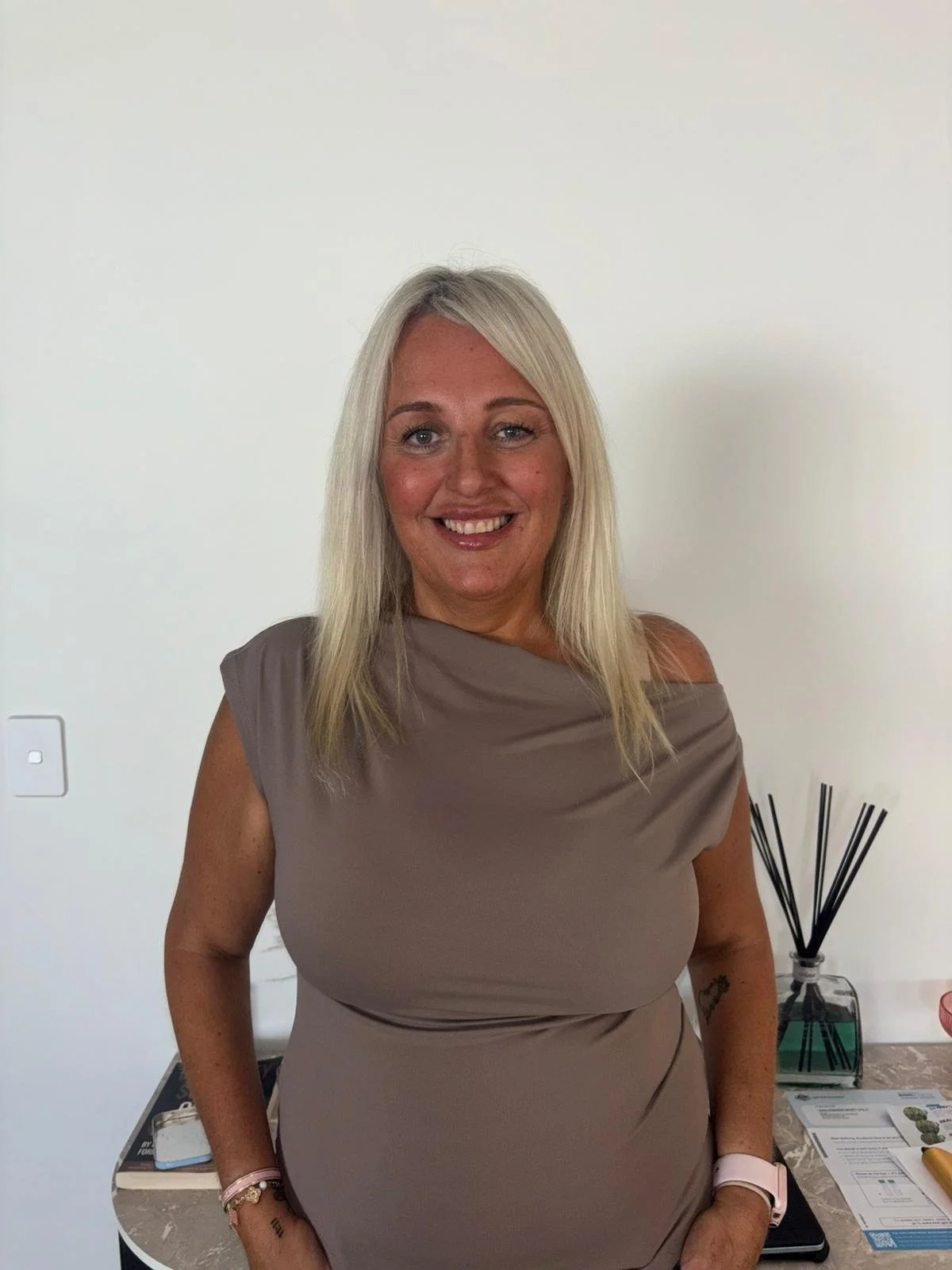 A smiling blonde woman in a taupe dress standing near a table with papers and a diffuser, against a plain white wall.