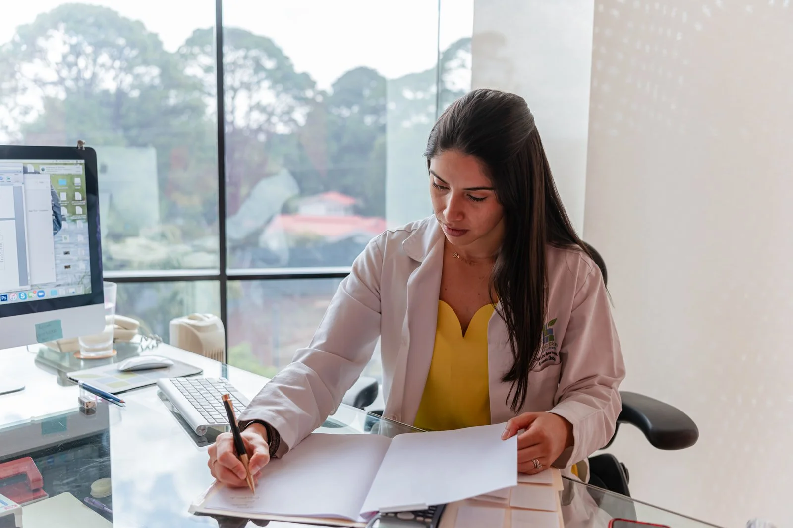 A woman working at a desk in an office, writing on papers with a pen. She is wearing a white coat over a yellow blouse, and there's a computer monitor, keyboard, and other office supplies on the desk.