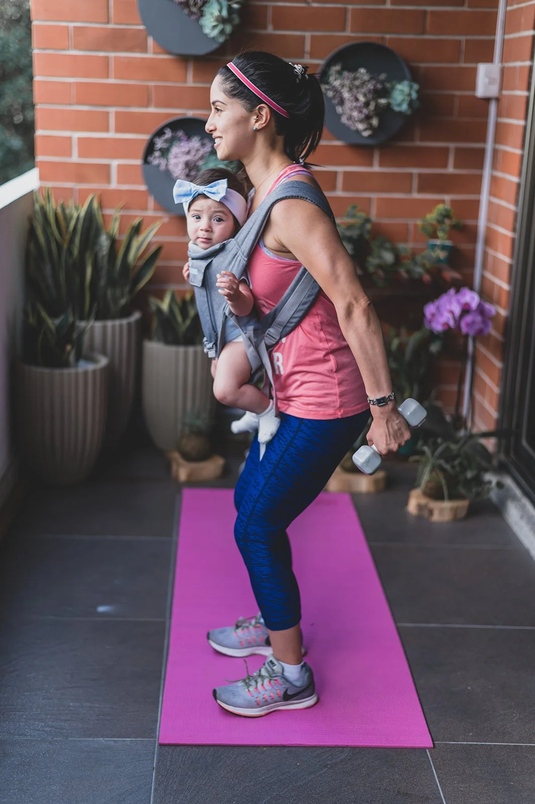 A woman is doing a workout with dumbbells while carrying a baby in a baby carrier on her chest on a purple yoga mat on a balcony with plants and a brick wall.