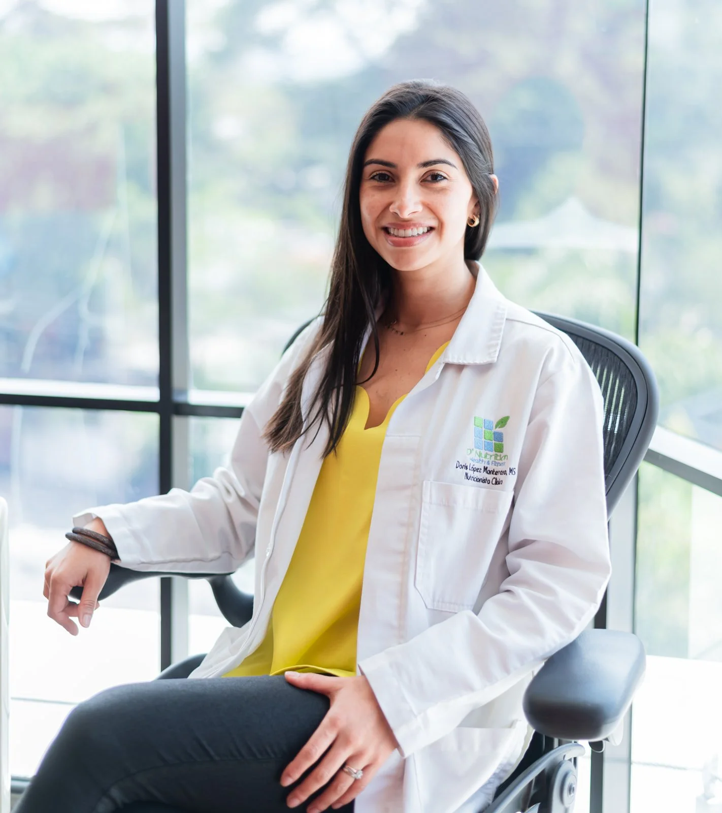A woman smiling, sitting in an office chair indoors with large windows in the background. She is wearing a white coat with a logo and text, a yellow top, and black pants. Doris Lopez Mgtr D´Nutrition Guatemala