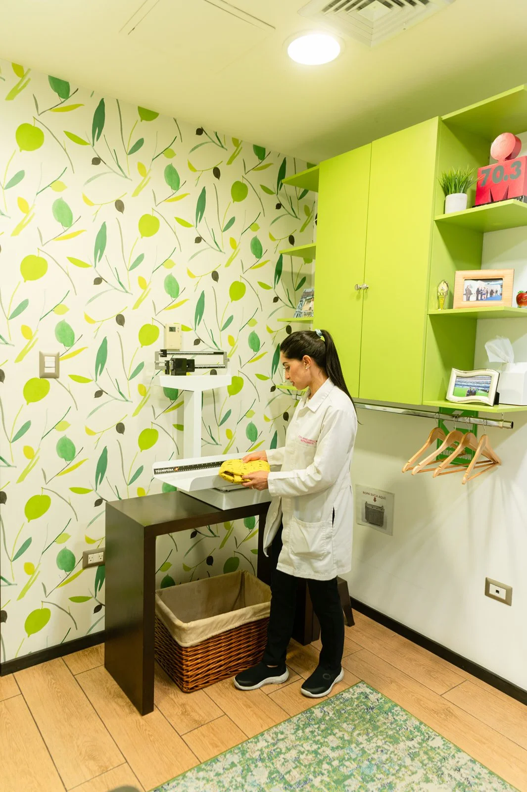 A woman in a white coat stands at a scale, weighing a yellow item in a brightly decorated room with green cabinets and wallpaper with green, yellow, and black abstract leaf and fruit patterns.