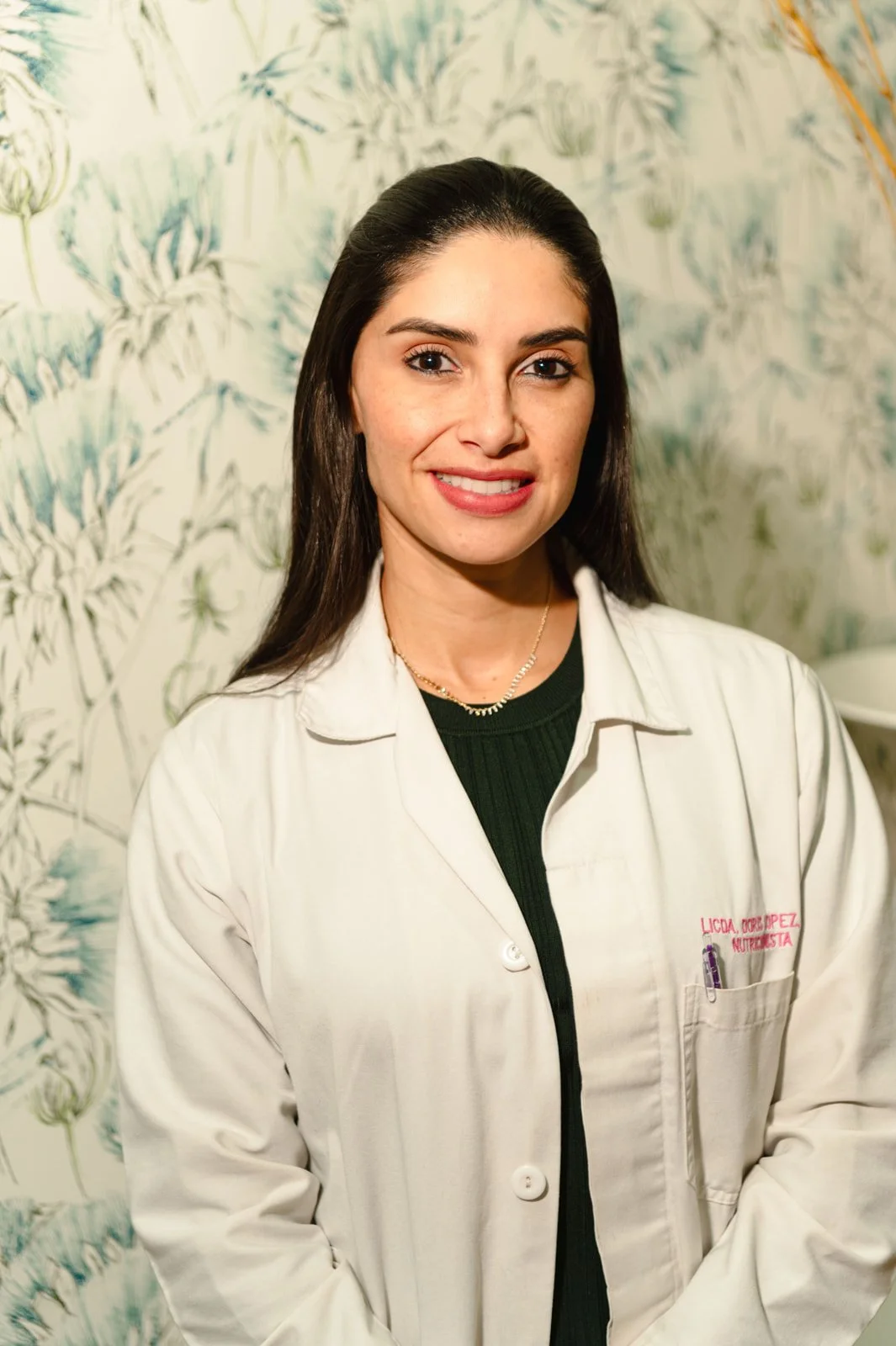 A young woman with long dark hair, smiling, wearing a white coat with embroidery and a black top underneath, standing in front of a leafy patterned background.