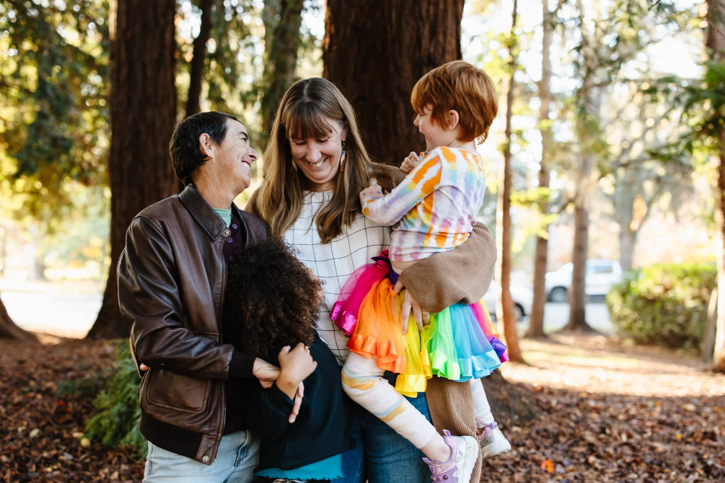 Three people, two adults and a young girl, are smiling and embracing each other outdoors in a wooded area, with sunlight filtering through the trees. The girl is wearing a rainbow-colored tutu.