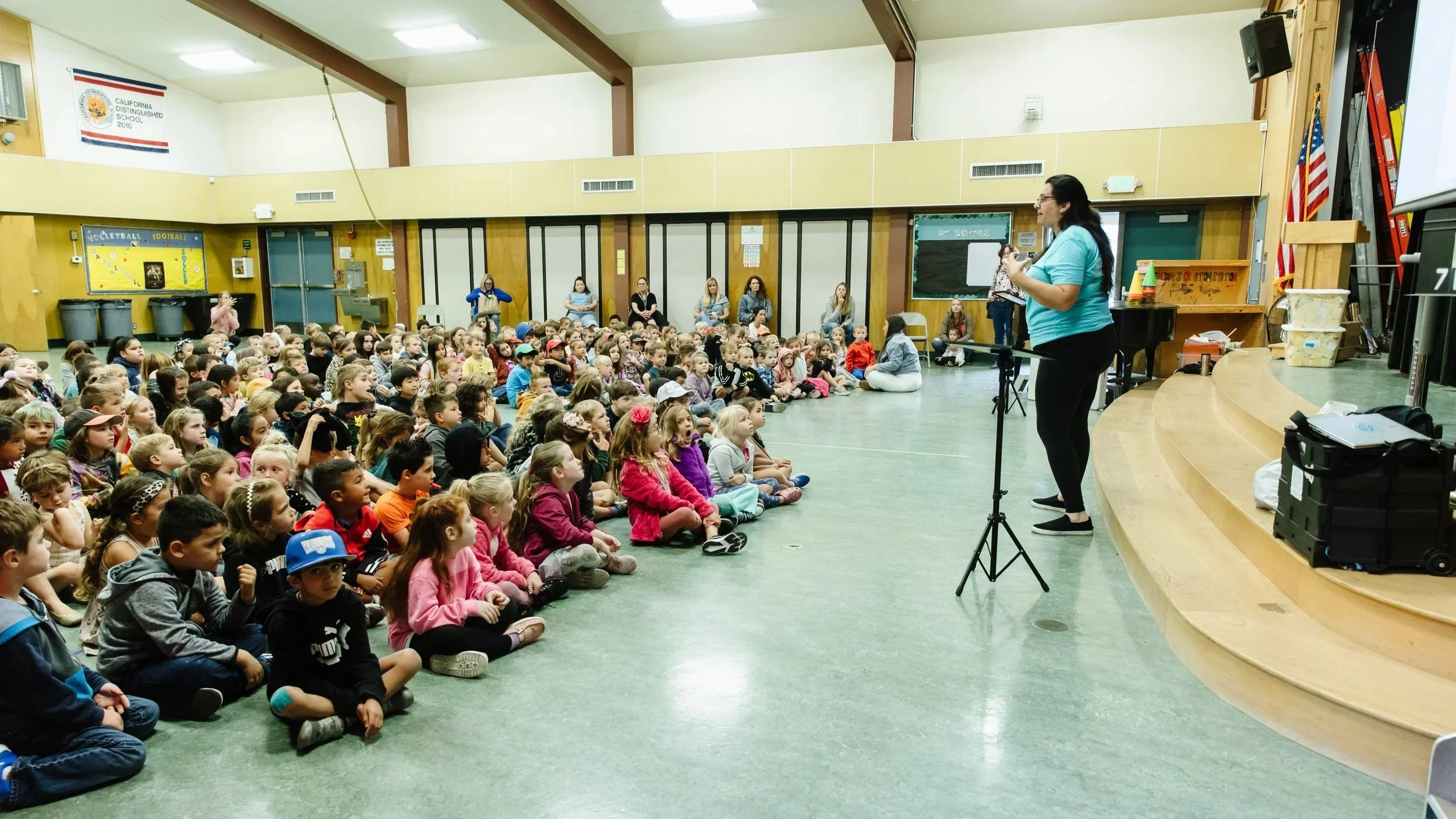 A large group of young children sitting on the floor in a school gymnasium, facing a woman who is speaking or performing with a musical instrument on a small stage. The room has beige walls, a California Distinguished School banner, and a blackboard with some children's artwork.