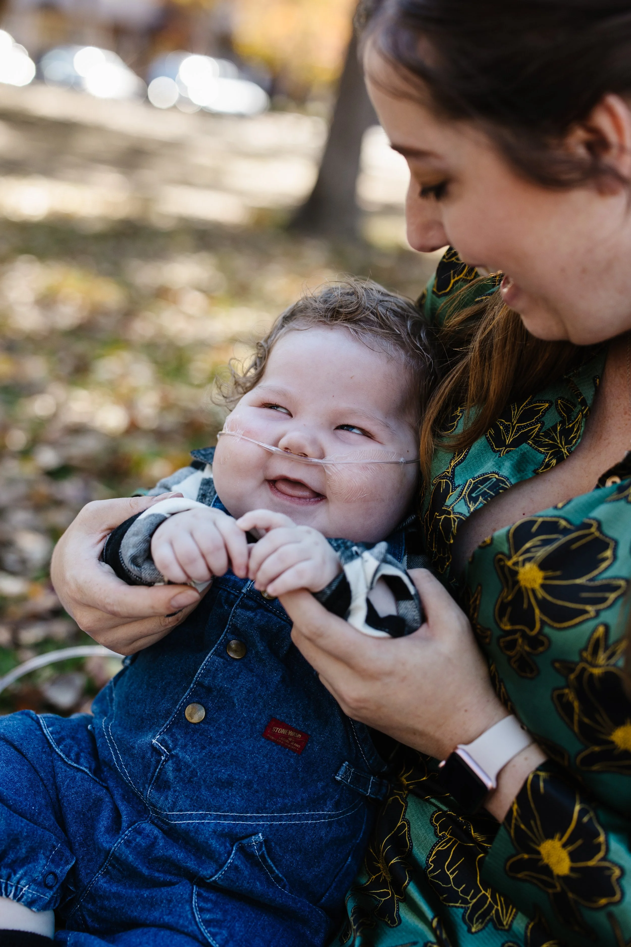 A woman holding a happy, smiling young child with a nasal cannula outdoors in a park with fall foliage.