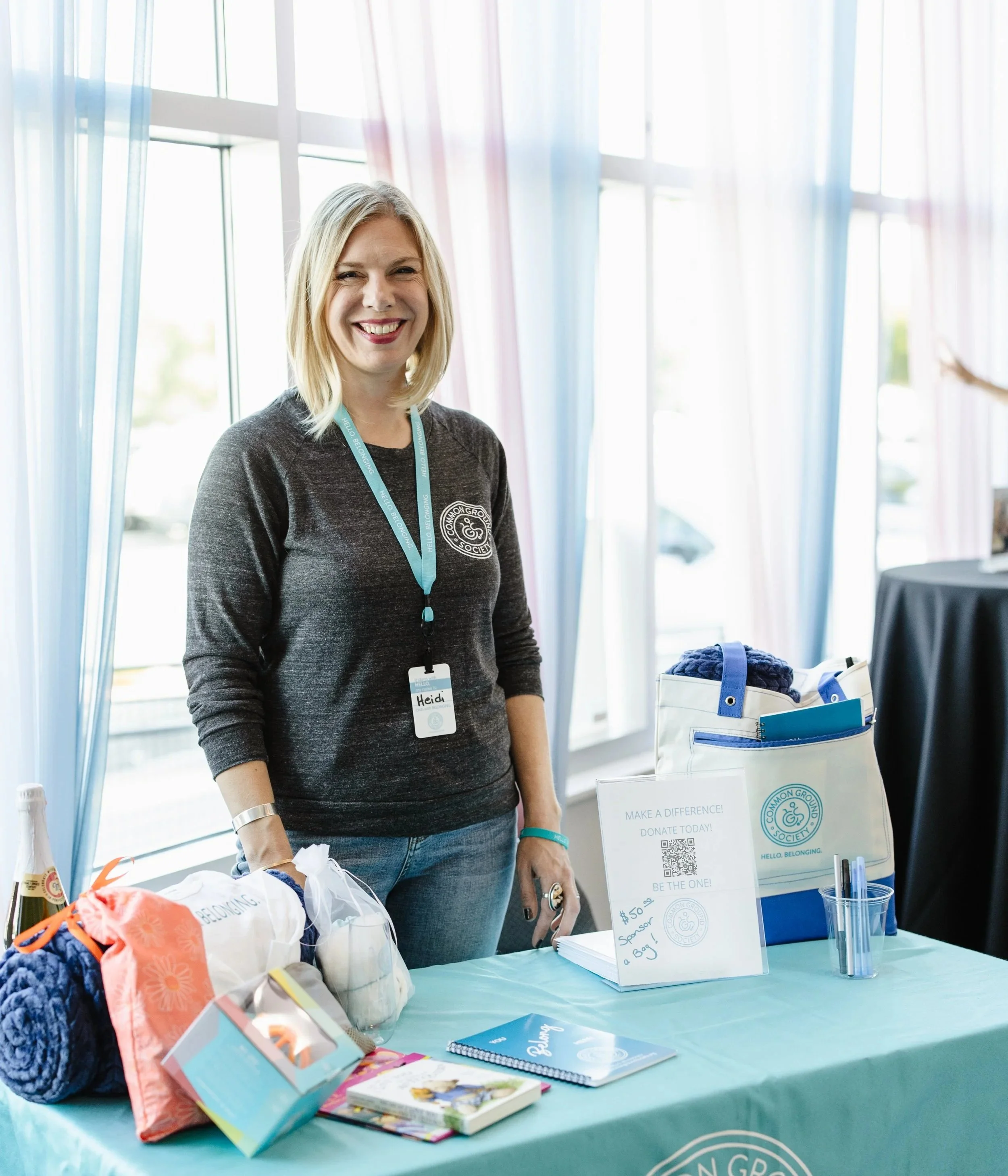 A smiling woman wearing a gray long sleeve shirt with a name tag at a table with charity fundraising items, including yarn, booklets, and donation sign, inside a room with large windows and light blue curtains.