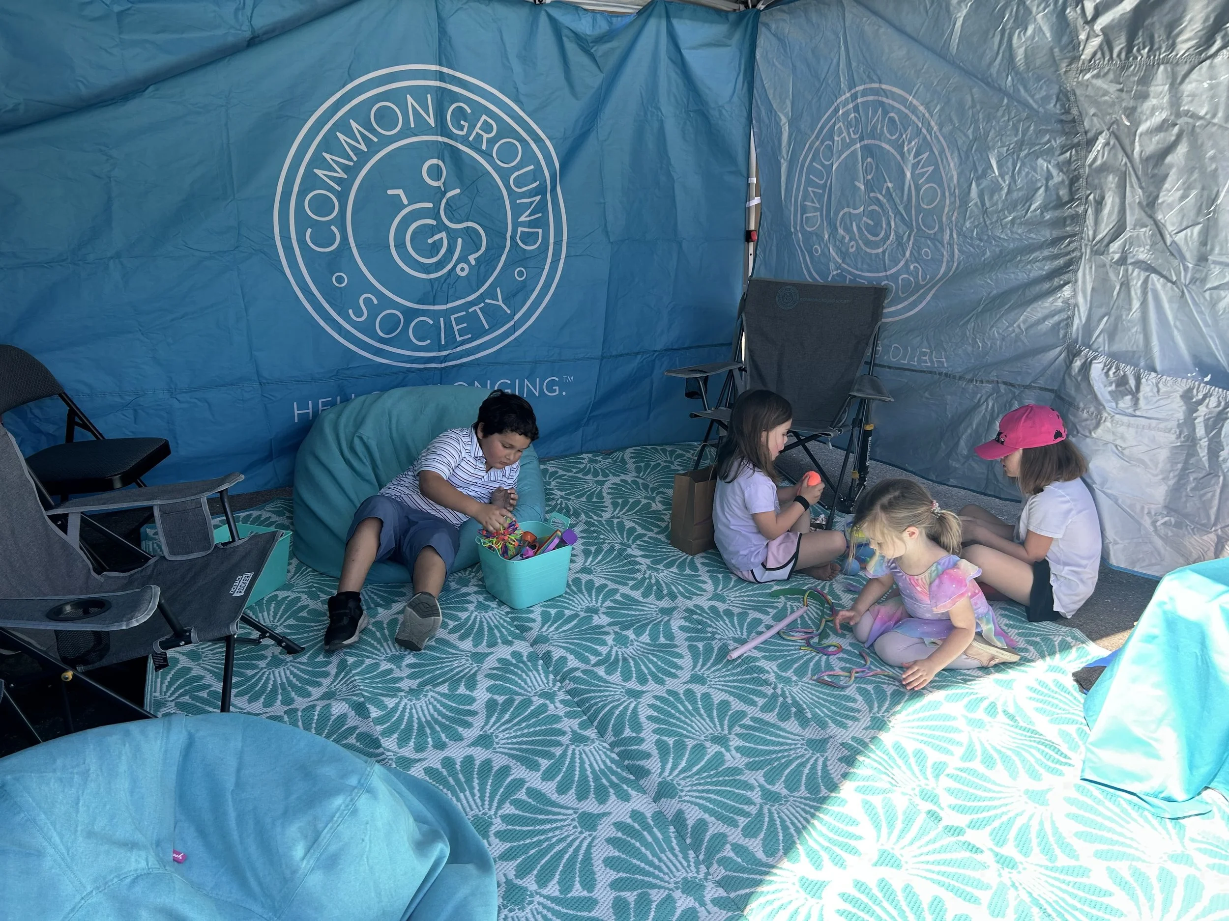 Children sitting on a patterned floor covering inside a blue tent with the Common Ground Society logo on the back wall, engaging in play activities.
