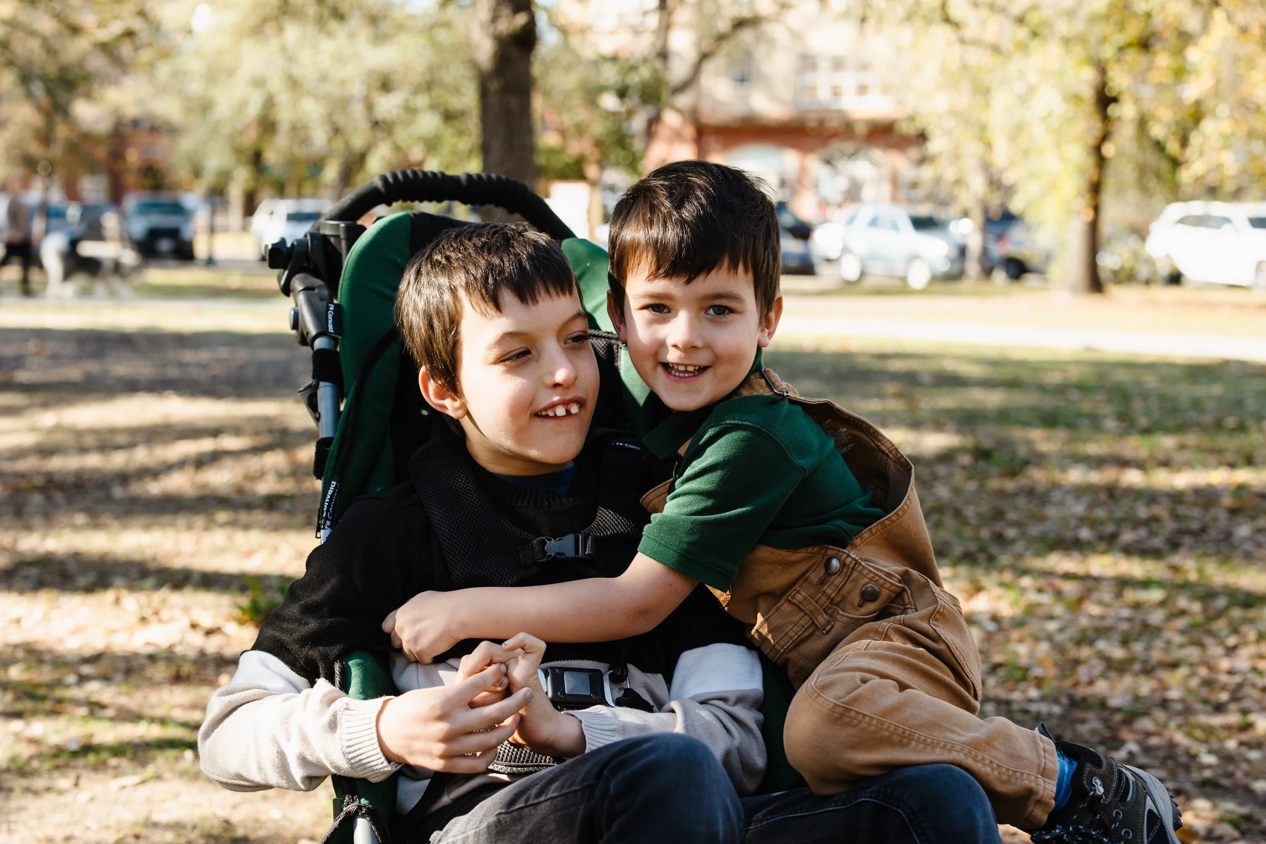 Two young boys with dark hair playing outside in a park on a sunny day, one sitting in a stroller and the other hugging him; trees and parked cars in the background.