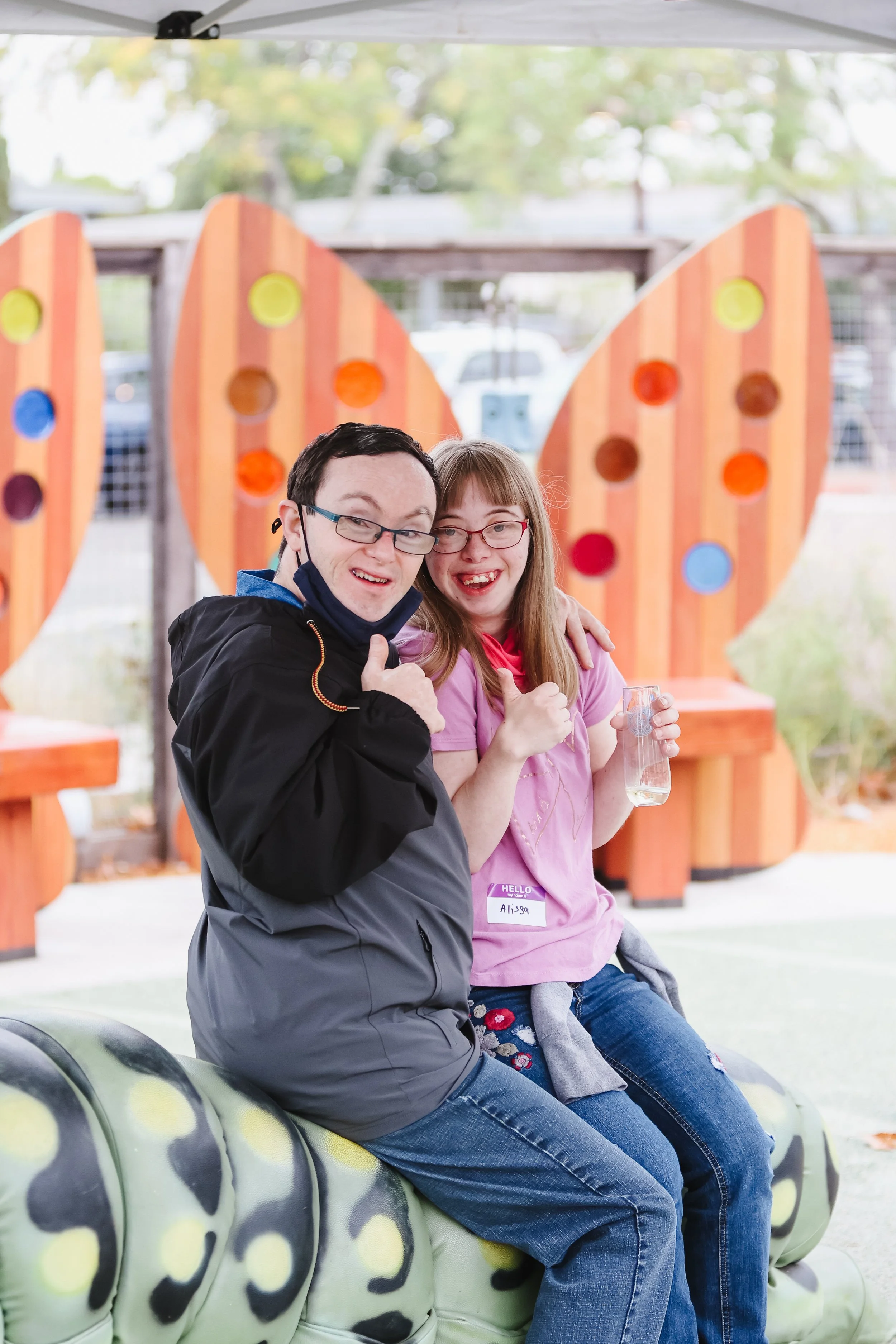 A man and a woman sitting on a decorated bench outside, smiling and giving thumbs-up. The woman is holding a drink. They are in front of a colorful wooden backdrop with circular cutouts at an event.