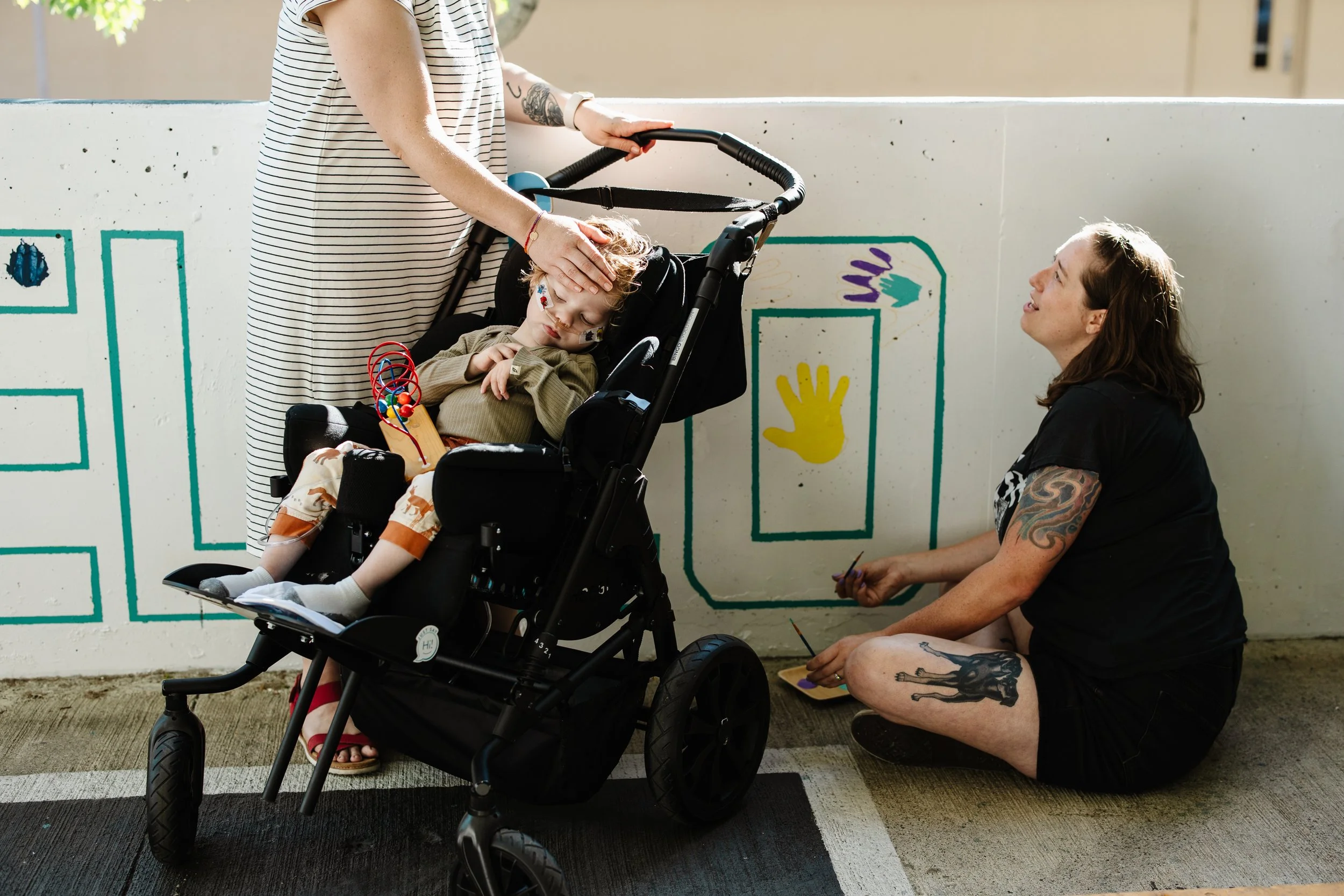 A woman with tattoos on her arm and leg sits on the ground painting colorful handprint art on a concrete wall. Another woman wearing a striped dress stands nearby, resting her hand on a child's head. The child is sitting in a stroller, sleeping, with face paint and a toy in hand, as the woman paints.
