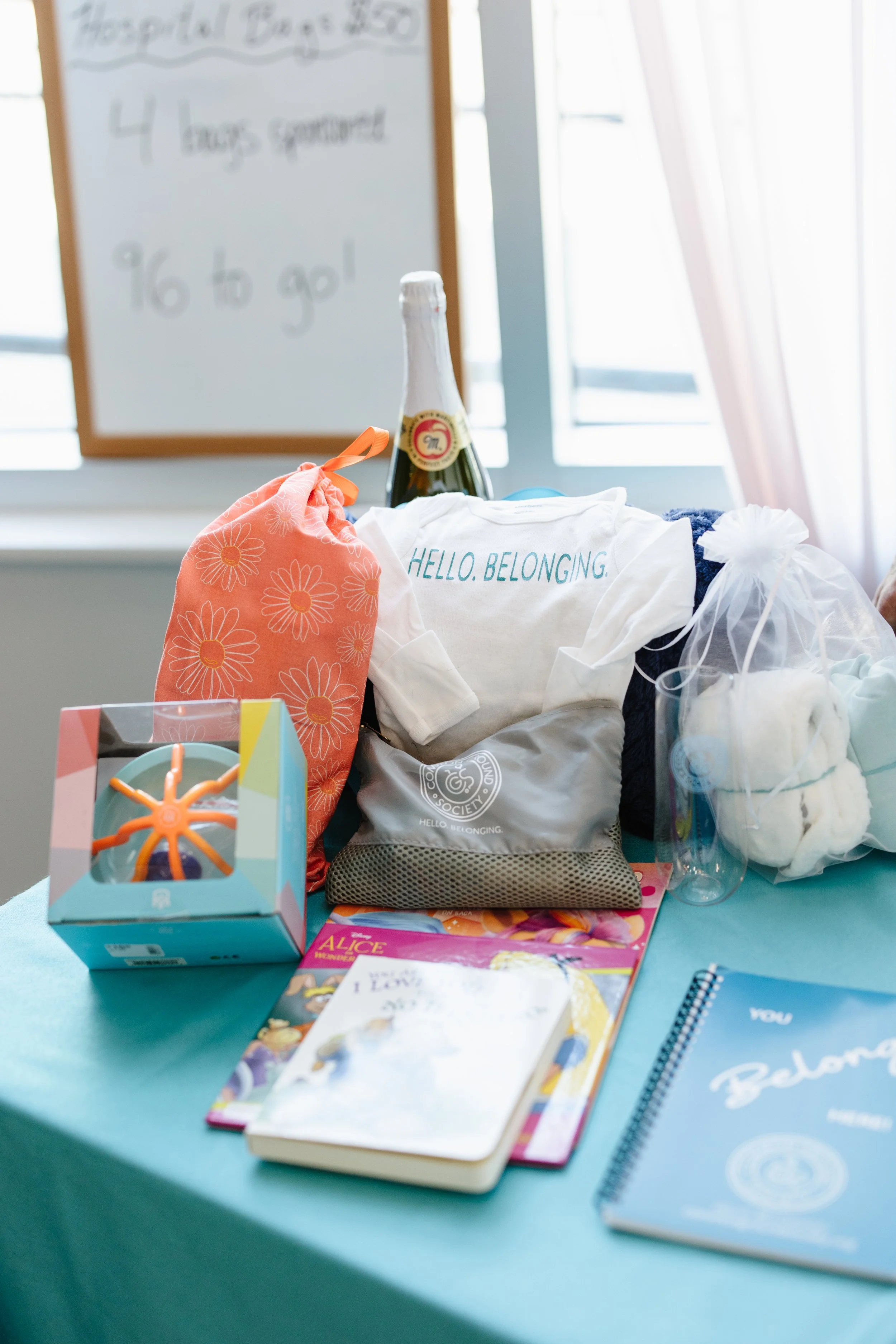 Table with welcome gifts including a white shirt with 'HELLO. BELONGING.' text, a boxed toy, books, a water bottle, a mesh bag with white items, and a gift bag with white fluff. In the background, a whiteboard with writing about a hospital class and a champagne bottle.