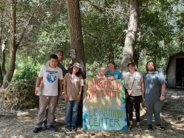 Group of seven people standing outdoors behind a colorful sign that reads 'Larkfield Community Learning Center,' surrounded by trees.