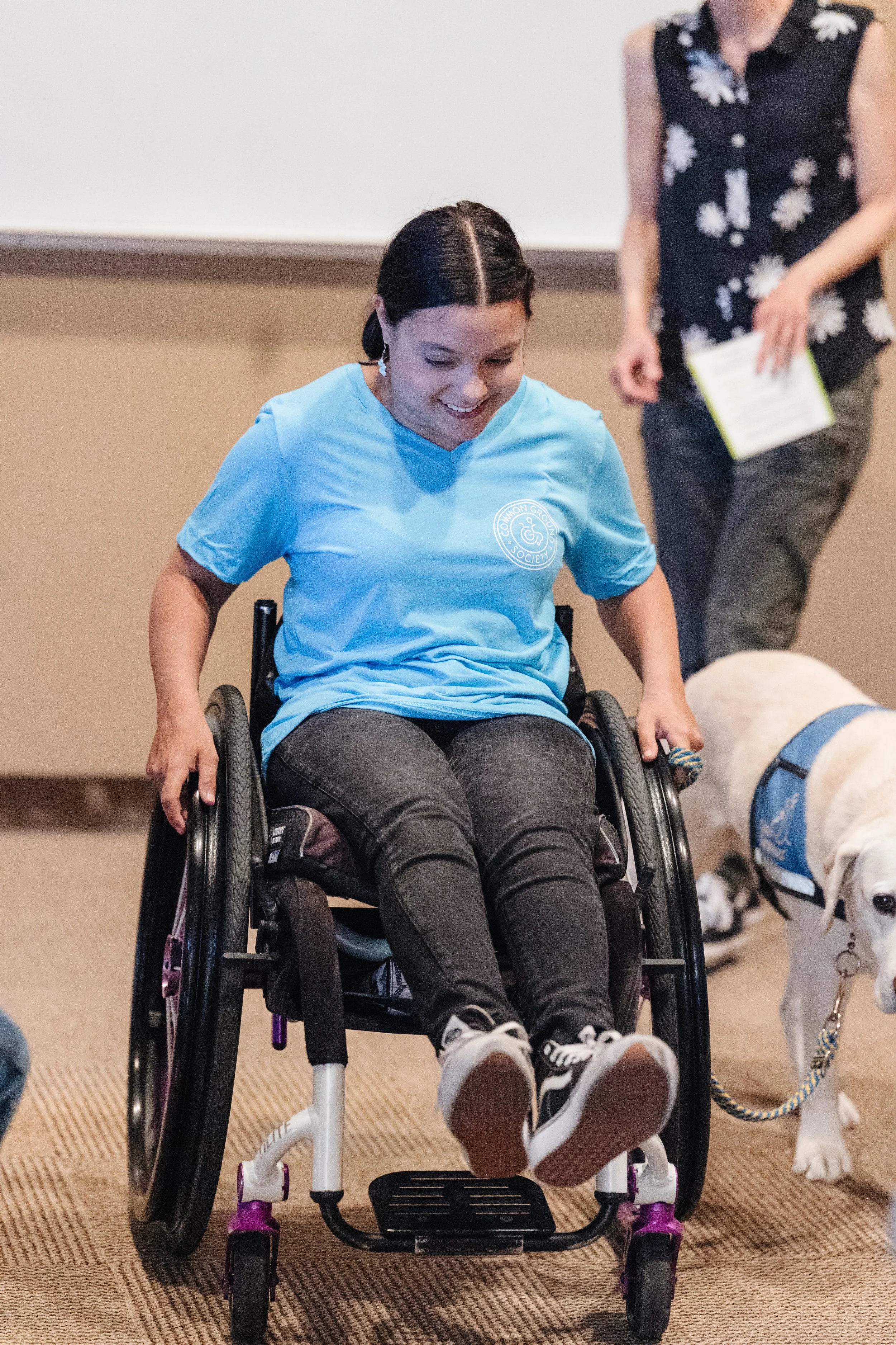 A young woman in a wheelchair smiling, with a service dog beside her, in an indoor setting.