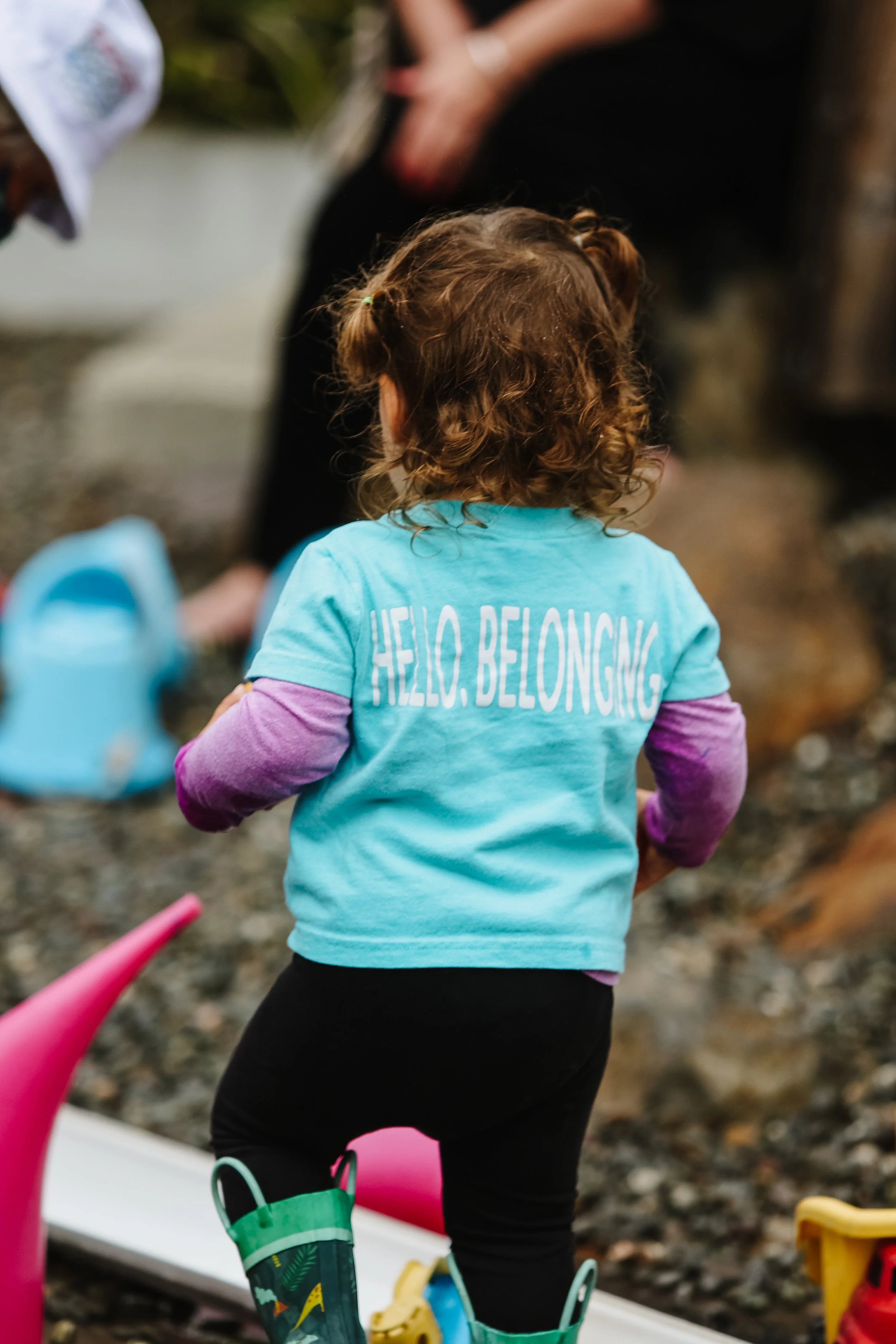 Back of a young girl with curly hair, wearing a blue shirt that says 'HELLO, BELONGING' and black pants, playing outdoors among toys on a gravel surface.