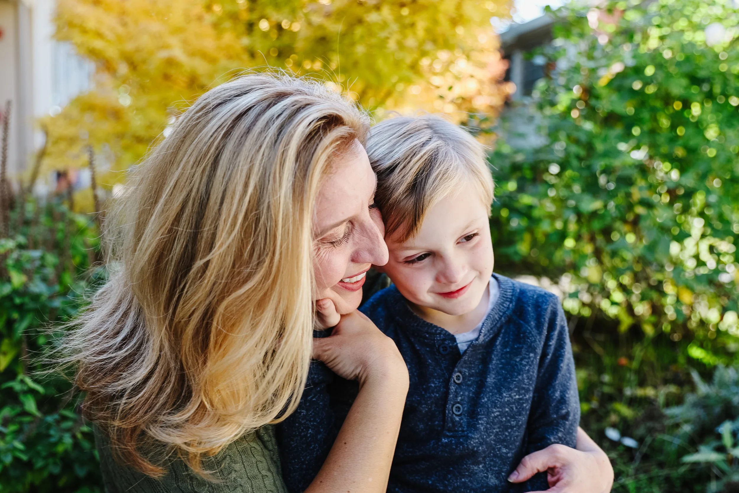A woman and a young boy smiling and hugging outdoors in a garden during autumn, with colorful fall foliage in the background.