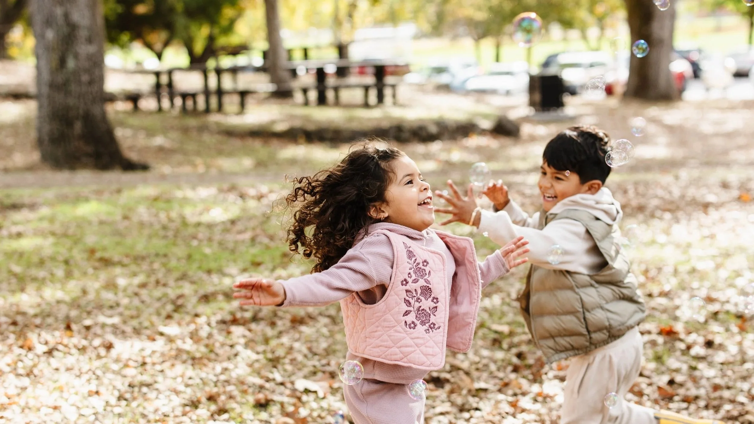 Two children, a girl and a boy, playing and laughing outdoors in a park surrounded by trees, with fallen leaves on the ground, and bubbles floating around them.